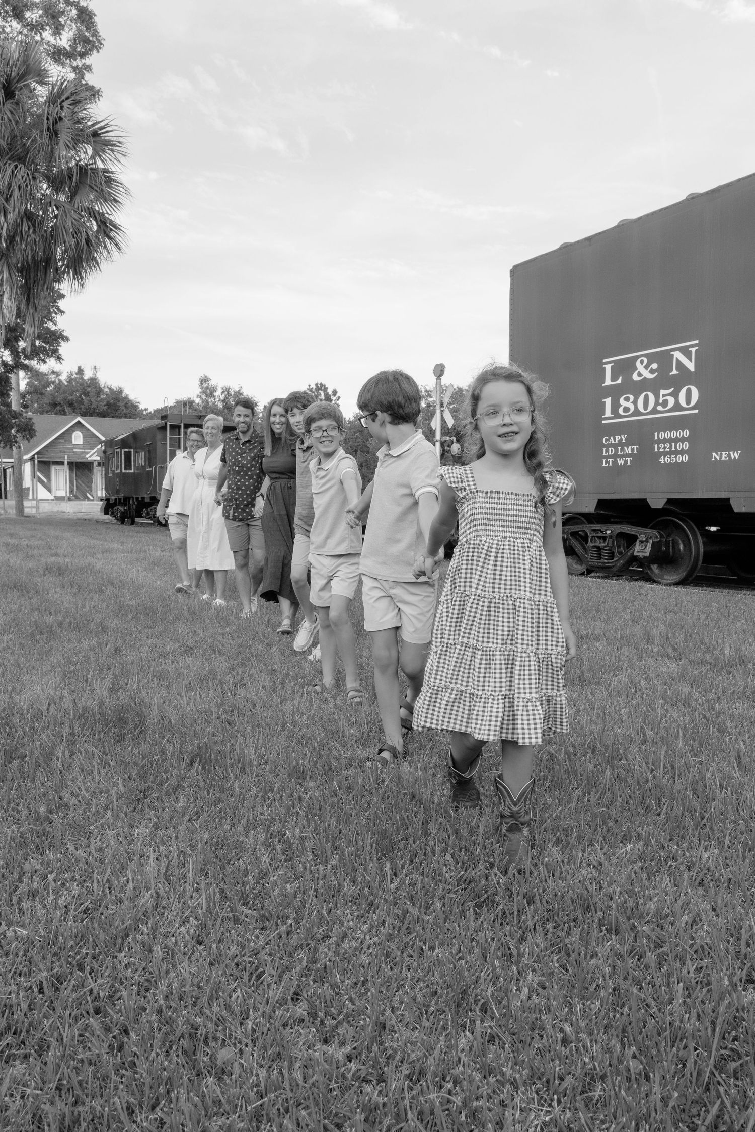 Children and adults walk in a line near an L&N railroad freight car 18050 on a grassy field in a vintage black and white photo.