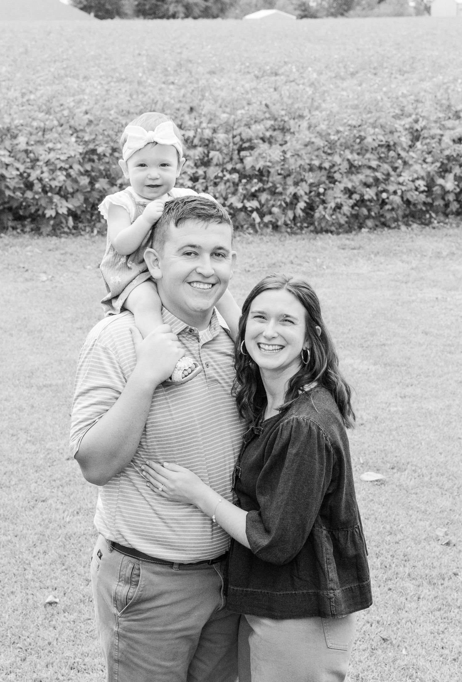 A black and white outdoor family portrait showing a couple and small child on shoulders in a garden setting.