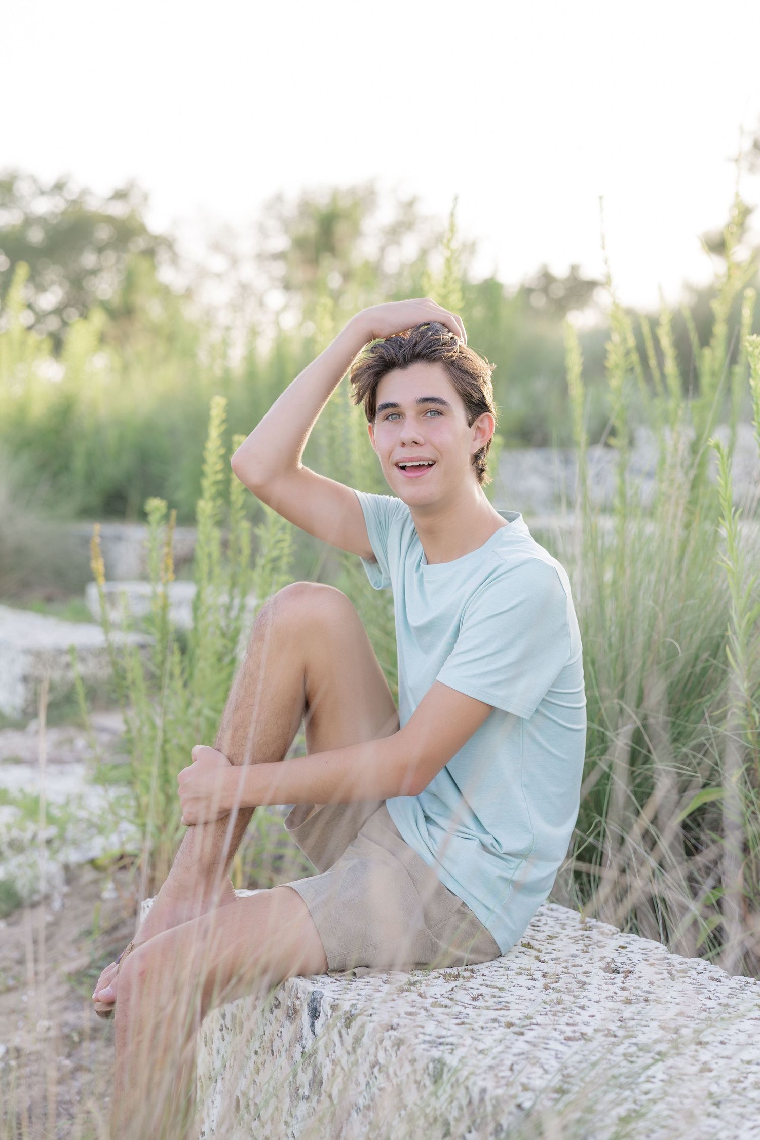 Young man in light blue t-shirt sitting on rocks outdoors, hand in hair, smiling, surrounded by tall green grass.