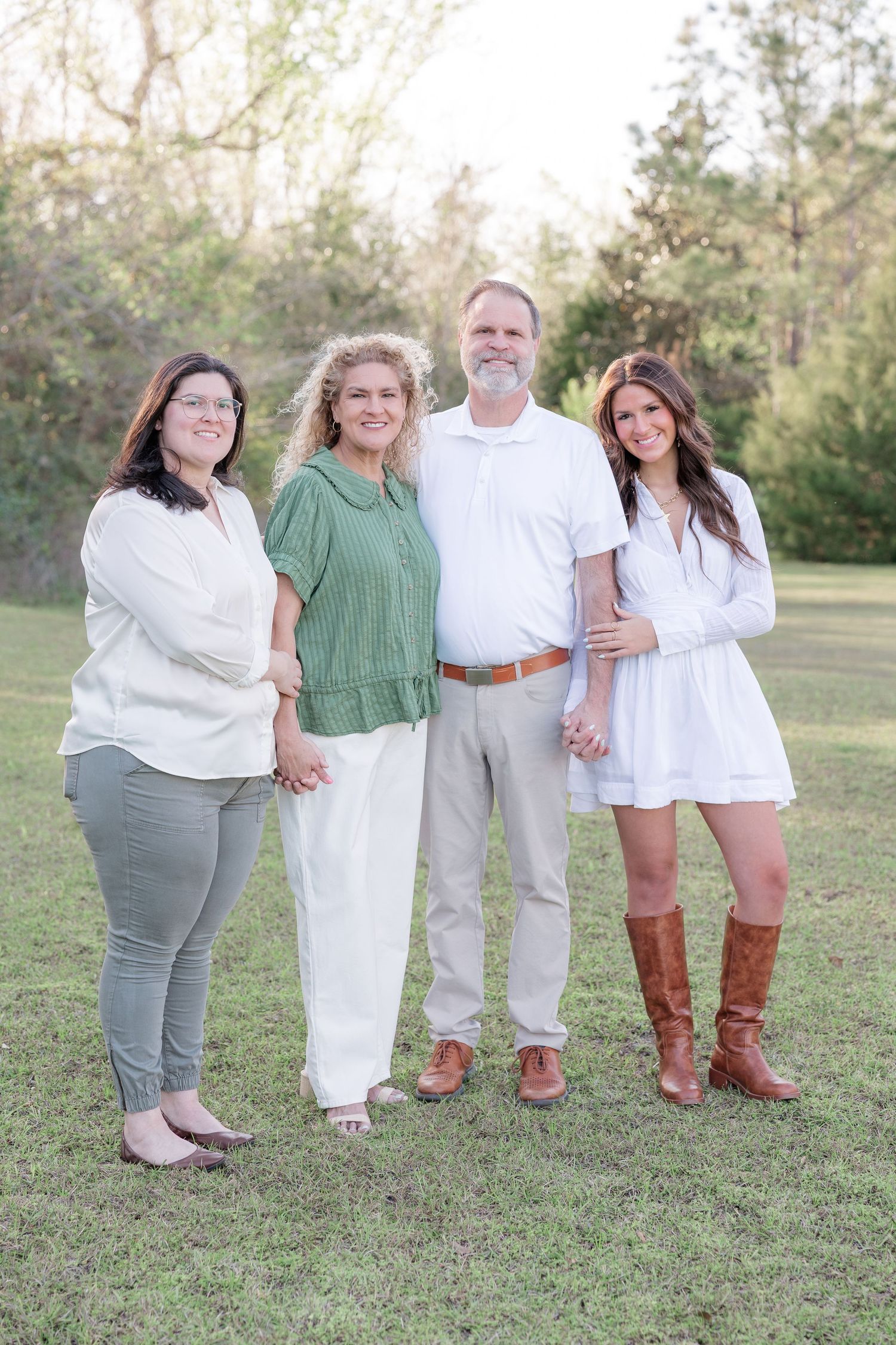 Family of four posing outdoors on green grass, wearing coordinated earth tones and white outfits, surrounded by trees.
