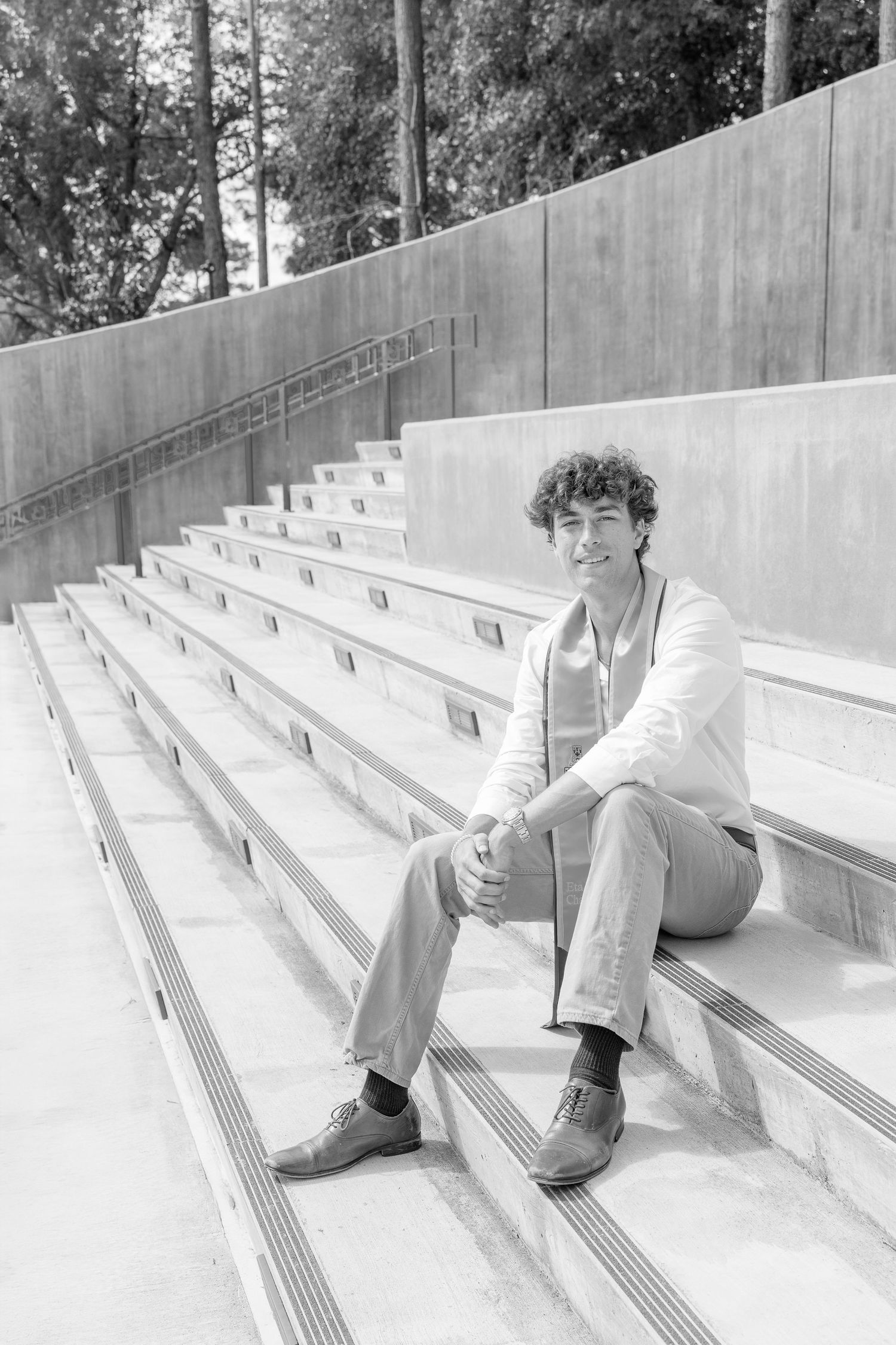 Young man with curly hair sitting on concrete bleachers outdoors, smiling, wearing light jacket and dress shoes.
