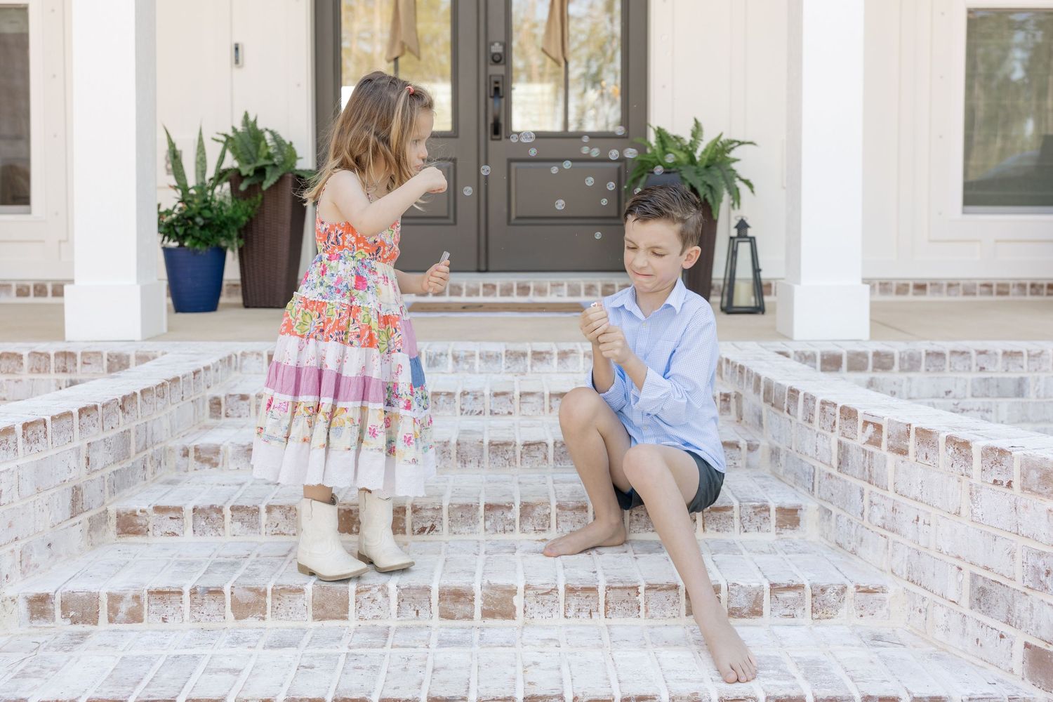 Two children playing with bubbles on brick porch steps, girl in pink dress and boy in blue shirt enjoying outdoor fun.