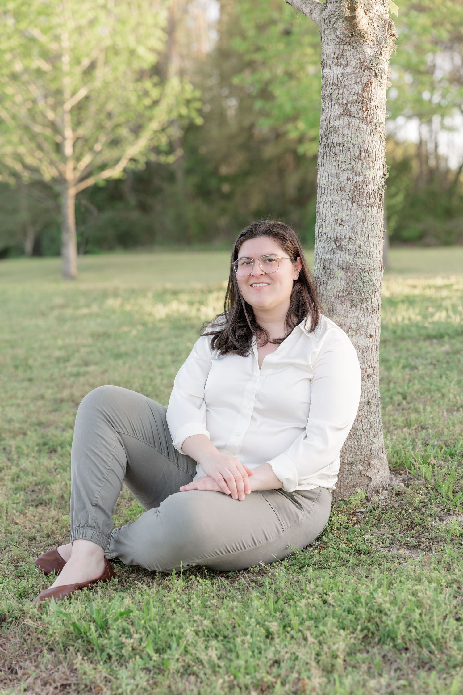 Woman in white blouse and gray pants sitting against a tree trunk on green grass in a sunny park.