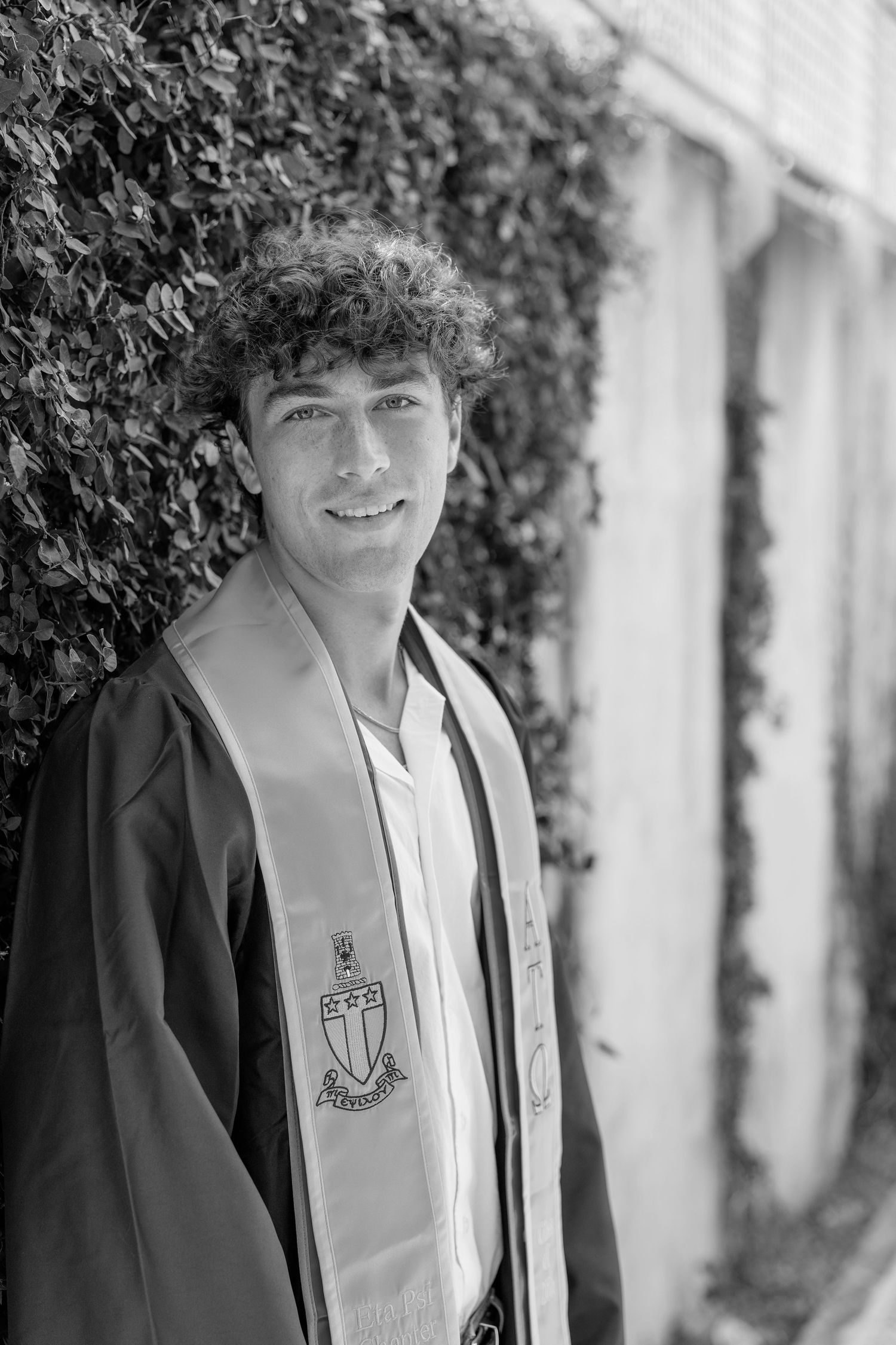 Young man in graduation gown and stole smiling outdoors against ivy-covered wall, black and white photo.