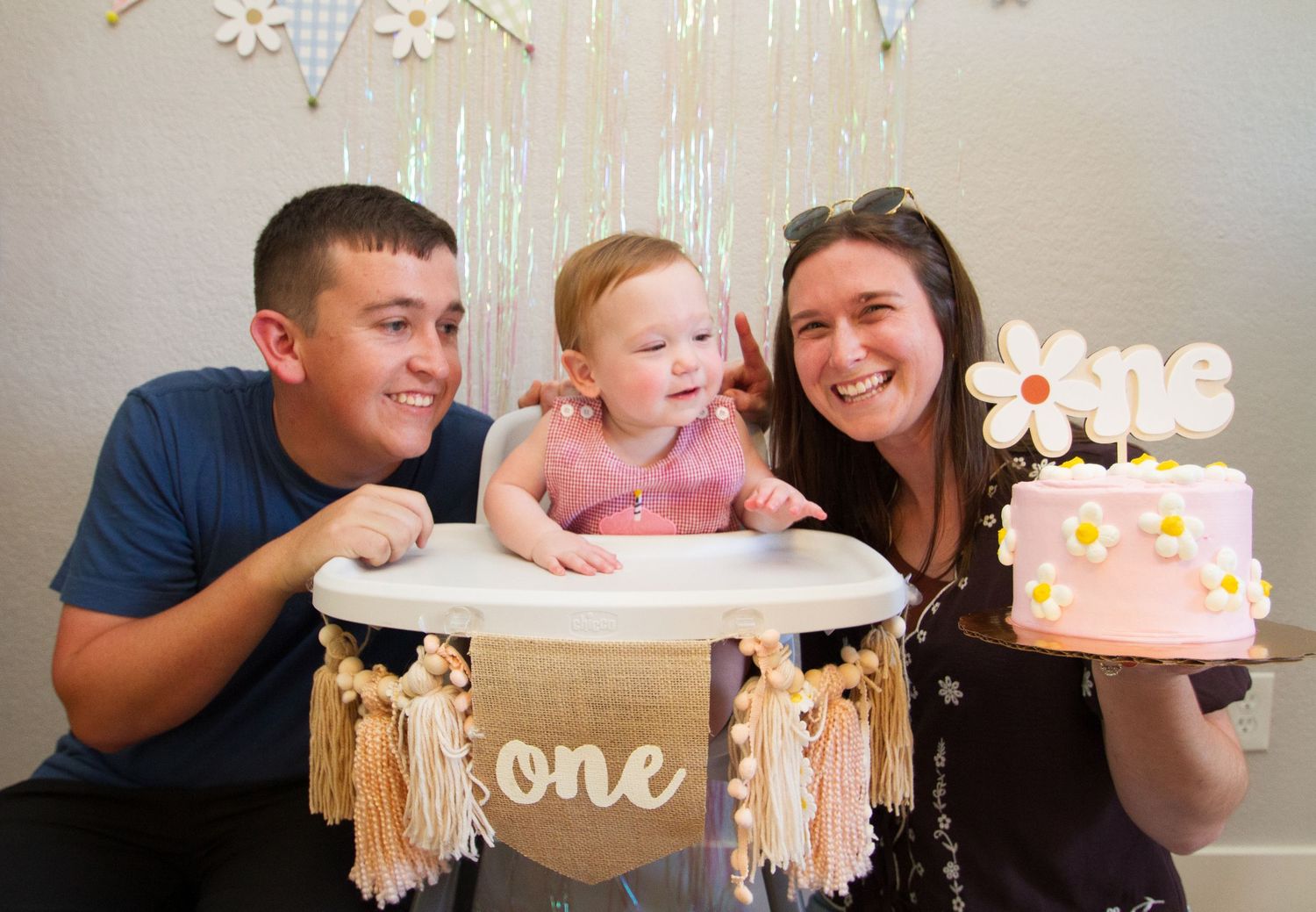 A baby sits in a high chair decorated with 'one' banner while someone holds a pink birthday cake with decorative flowers.