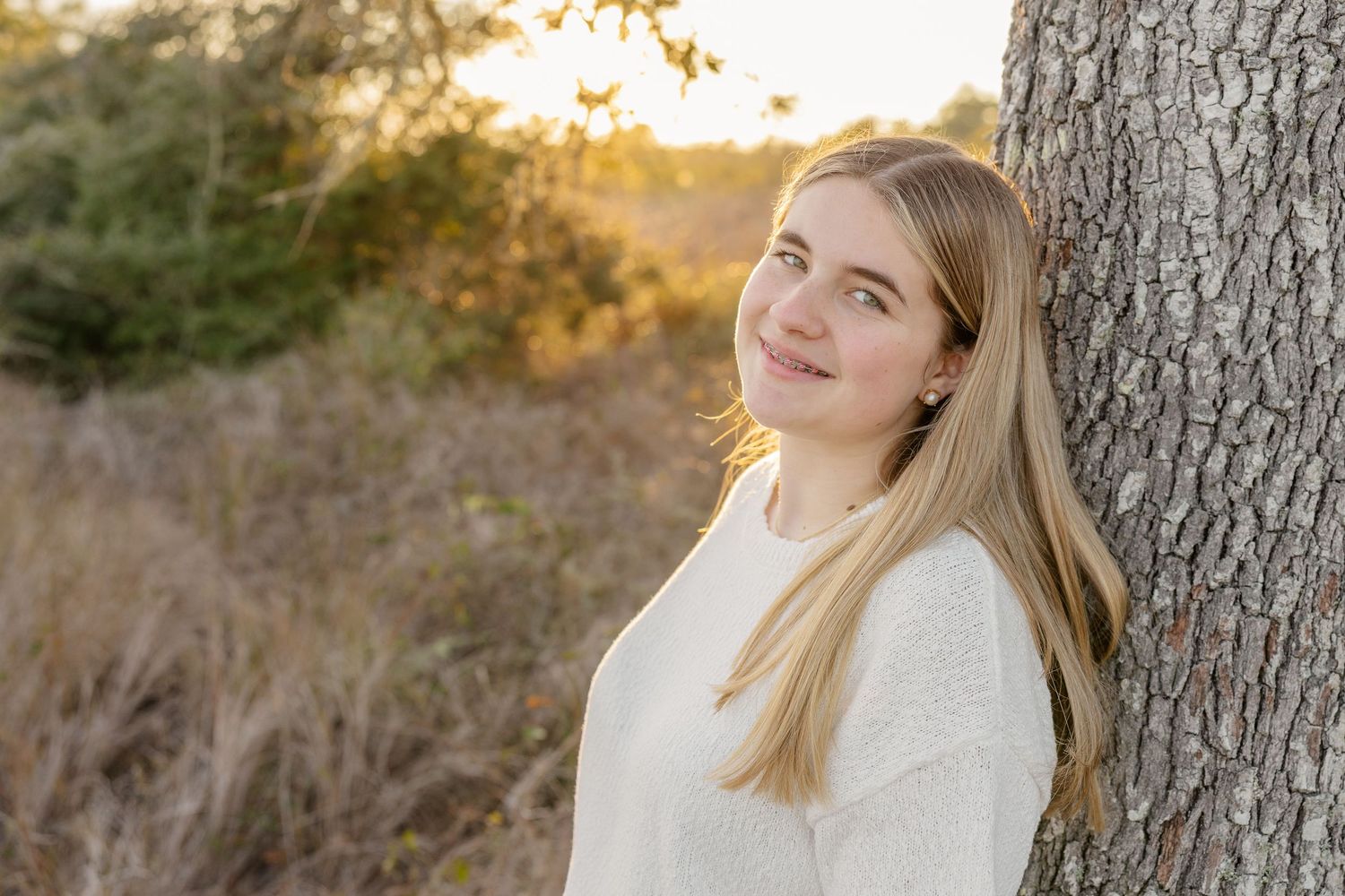 A woman with blonde hair poses against a tree trunk during golden hour in a natural outdoor setting.
