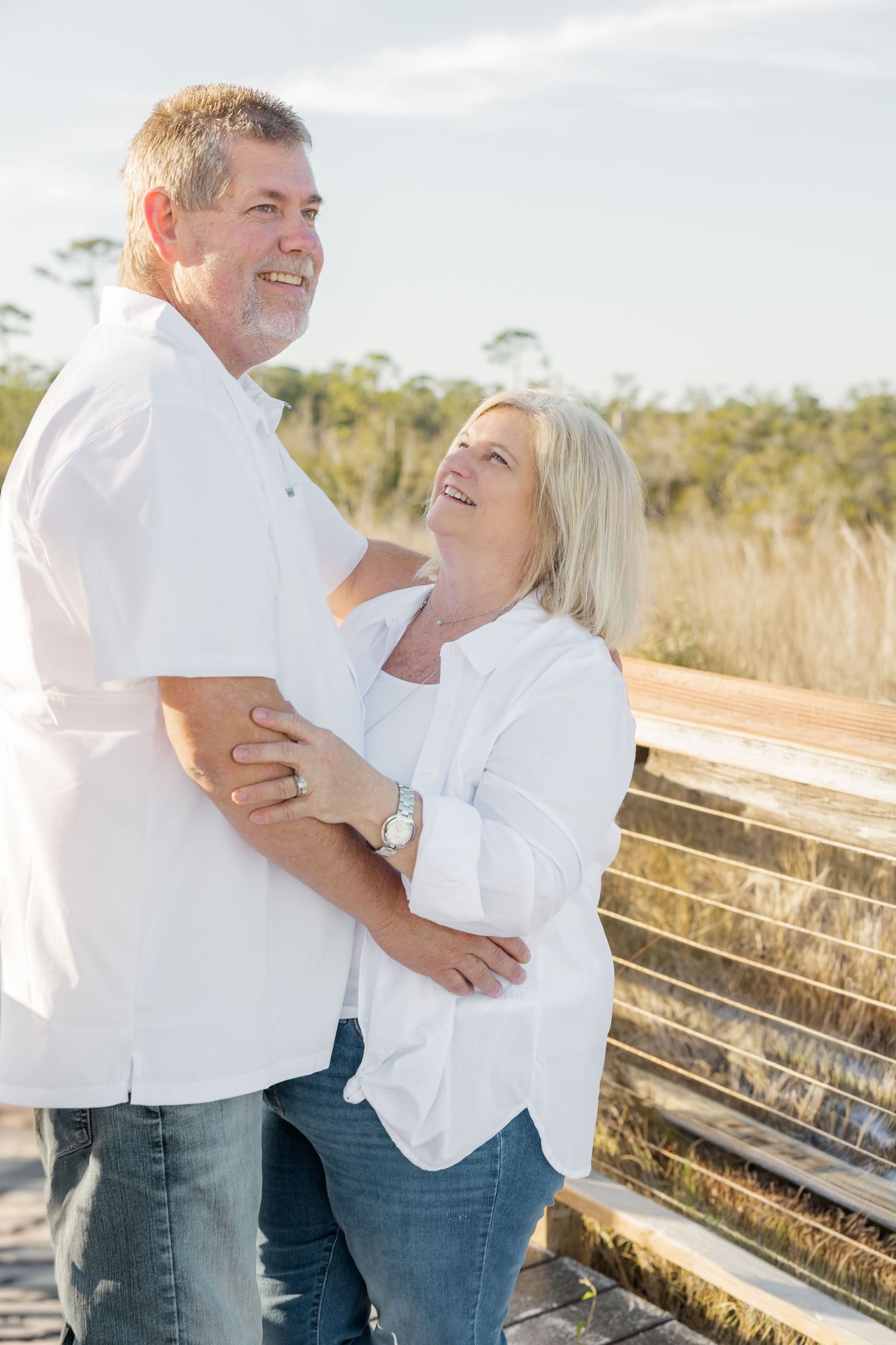 A couple in white shirts sharing joyful moments together on a wooden boardwalk during a sunny outdoor photoshoot.
