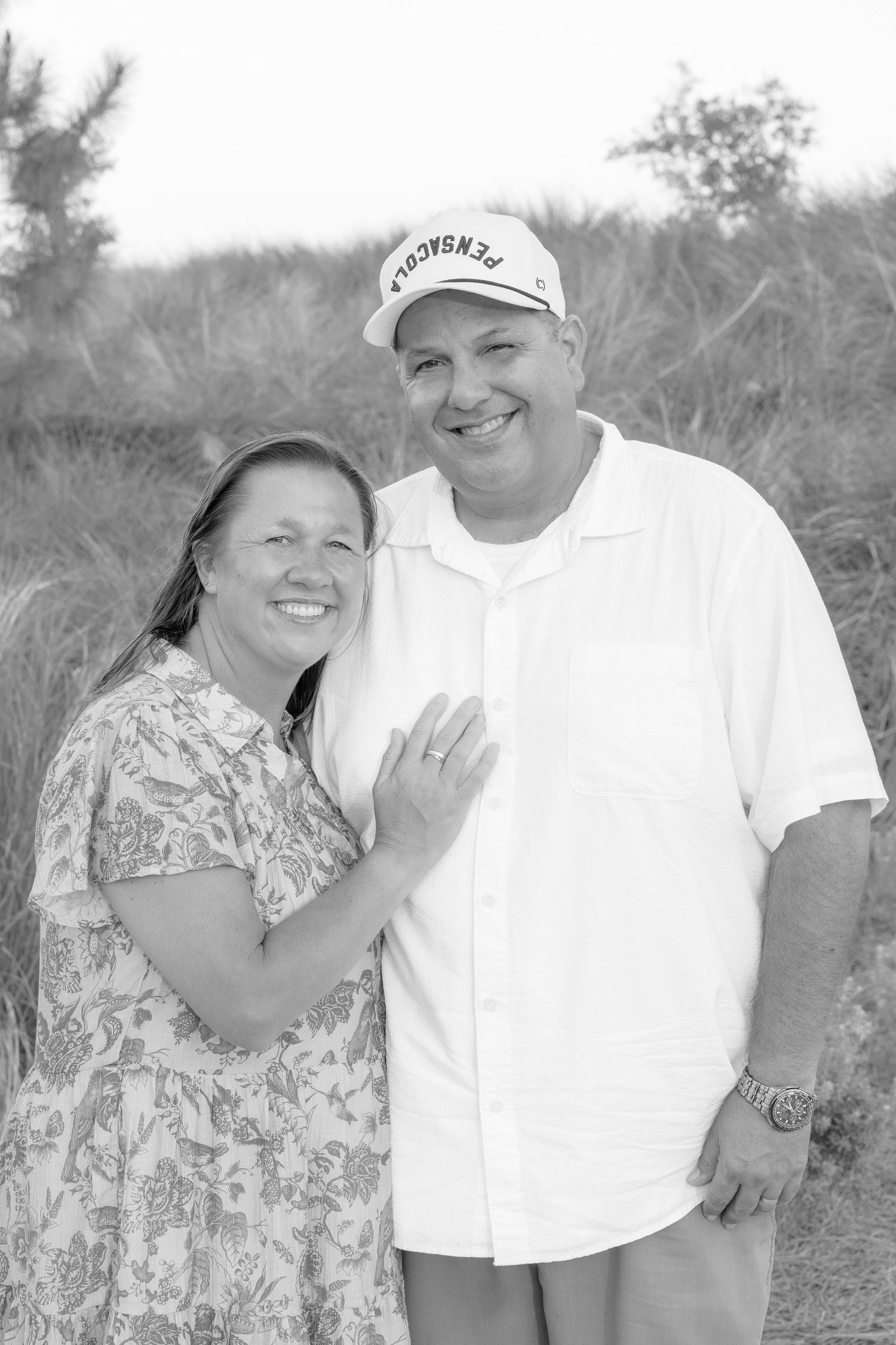 Black and white photo of a smiling couple outdoors, man in cap and white shirt, woman in floral dress, posing together.