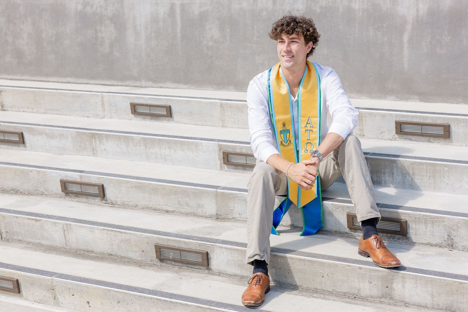 Smiling graduate sits on concrete steps wearing blue and gold graduation stole and white dress shirt.