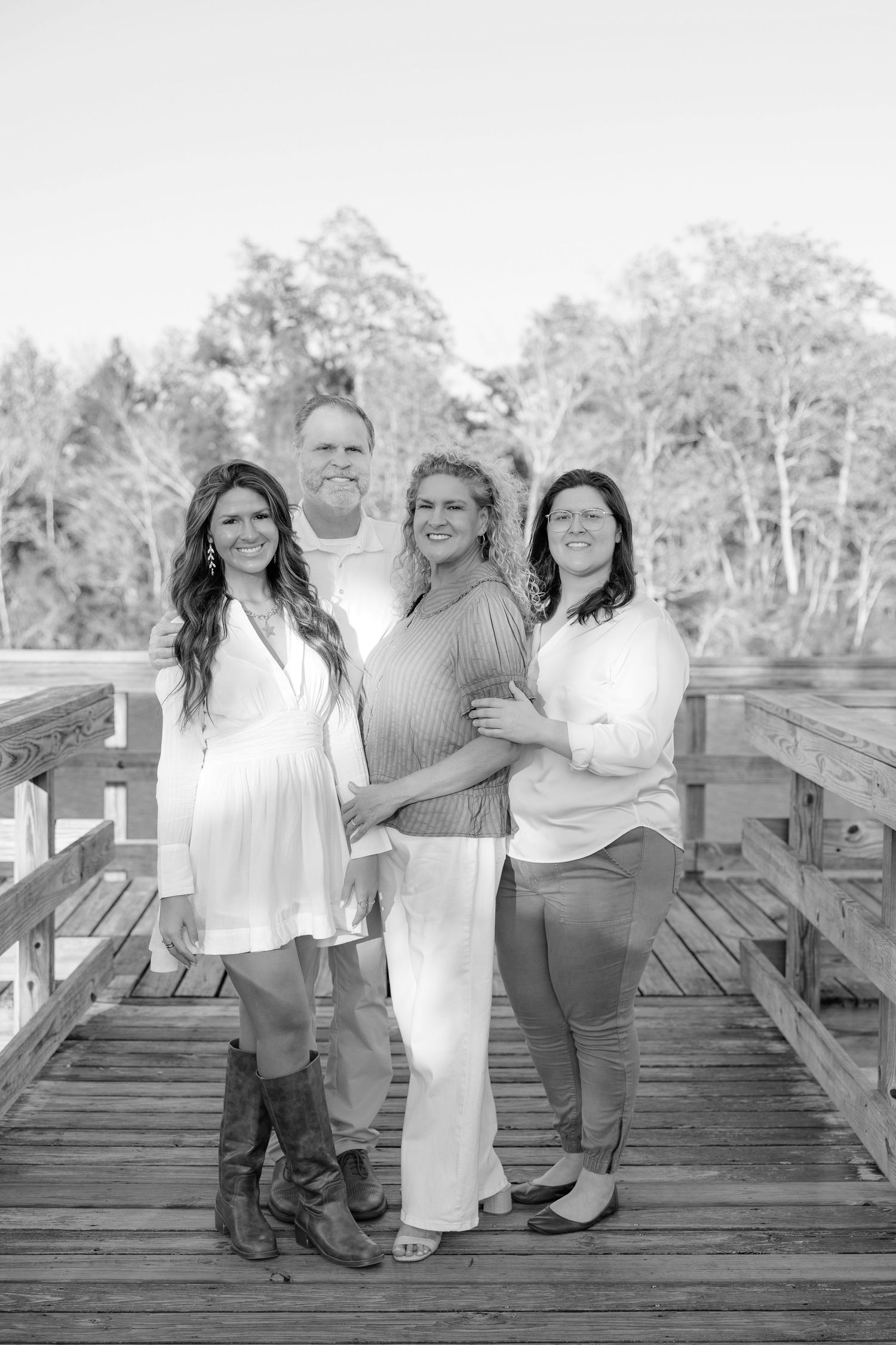 Black and white family portrait of four adults smiling on a wooden bridge surrounded by trees.