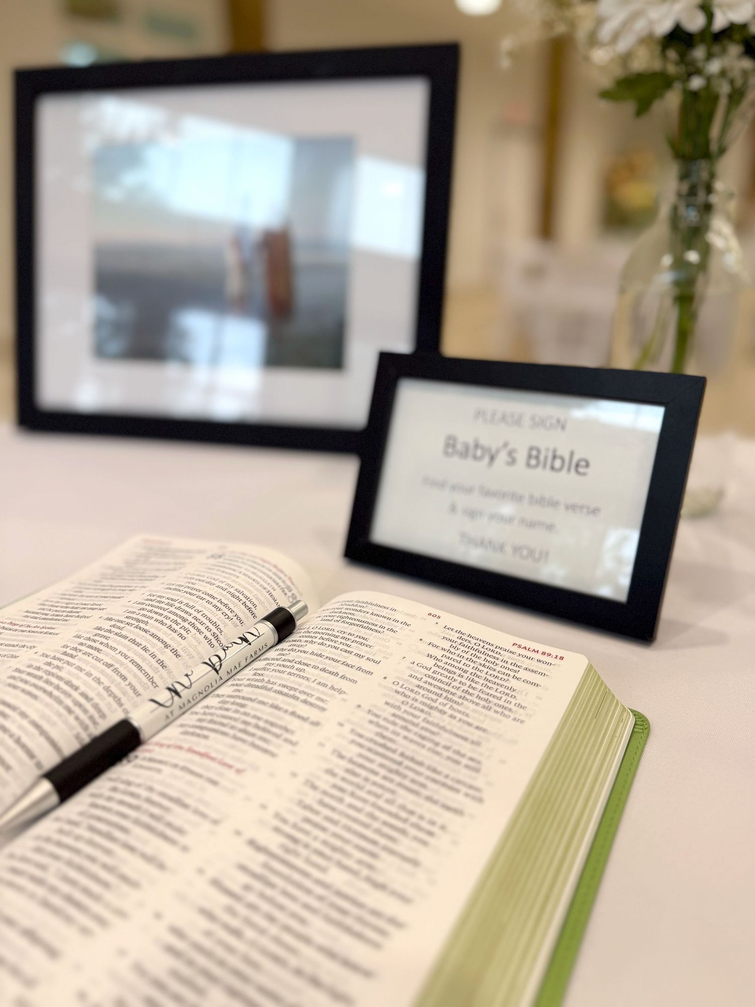 An open Bible displayed on a white table with flower arrangements and framed photos in the background.