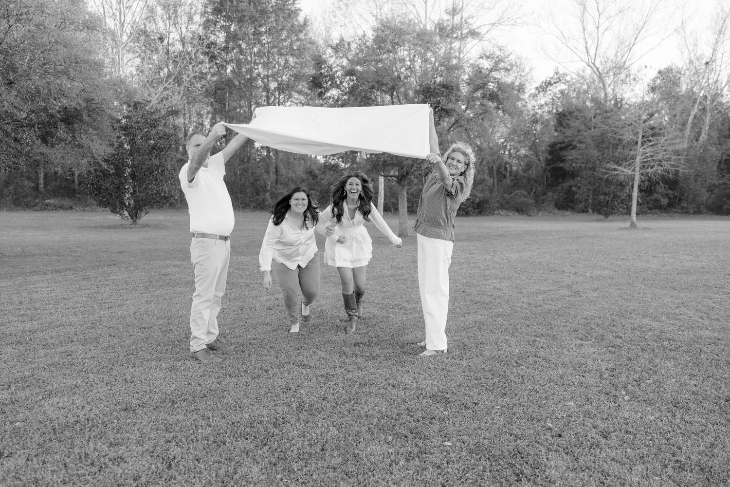Black and white photo of a family of four playing with a blanket outdoors in a grassy park setting.