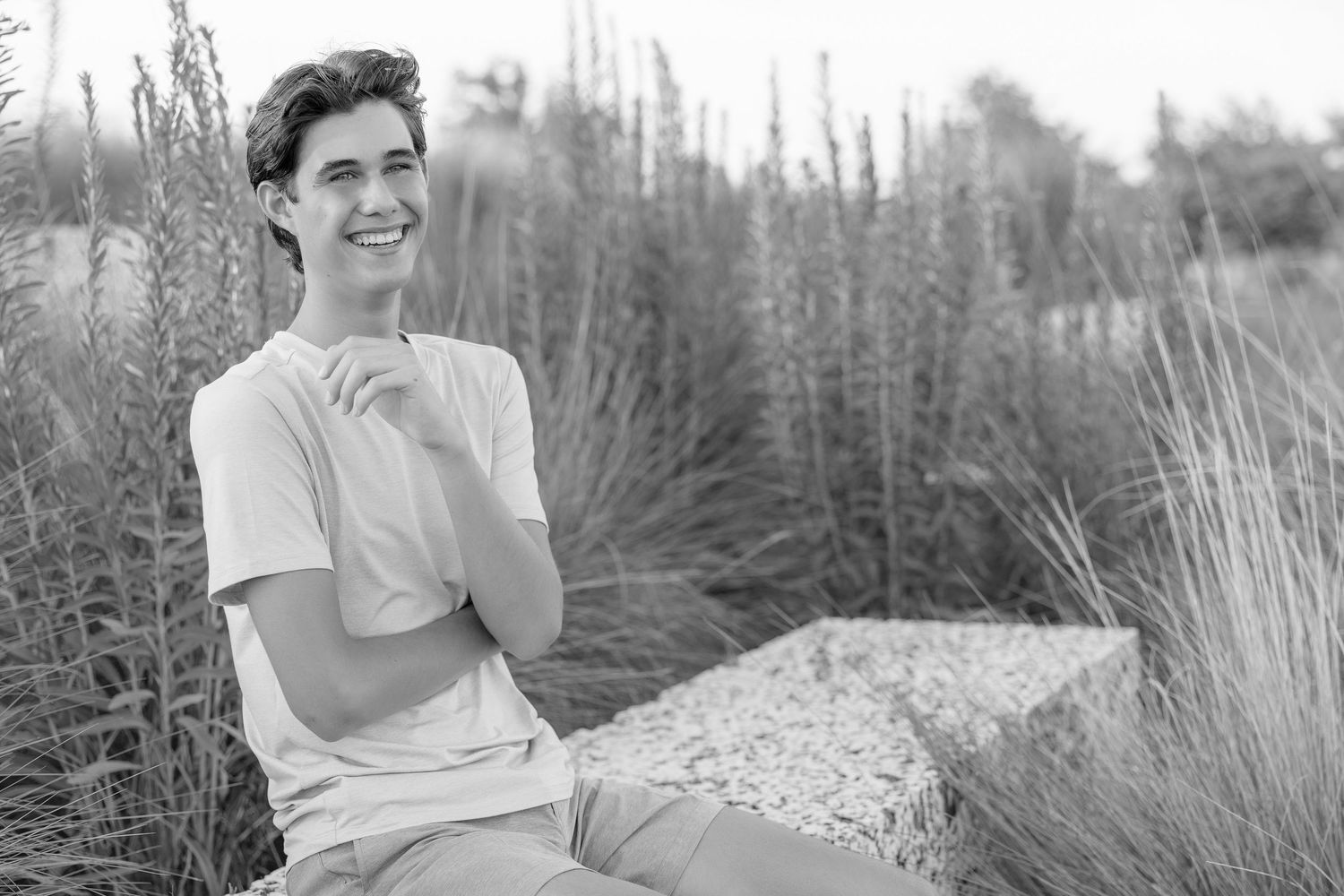 Smiling young man in white t-shirt sitting outdoors among tall ornamental grasses in a black and white photo.