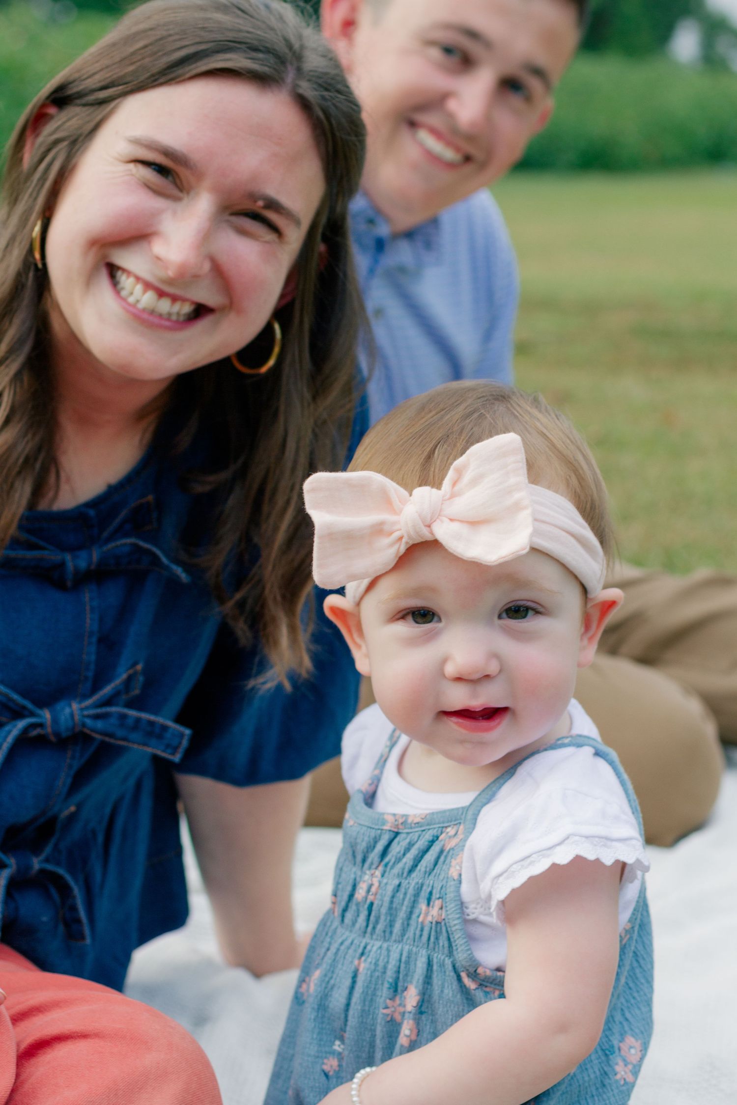 A family sits together outdoors on a sunny day, with a baby wearing a blue dress and pink bow headband.