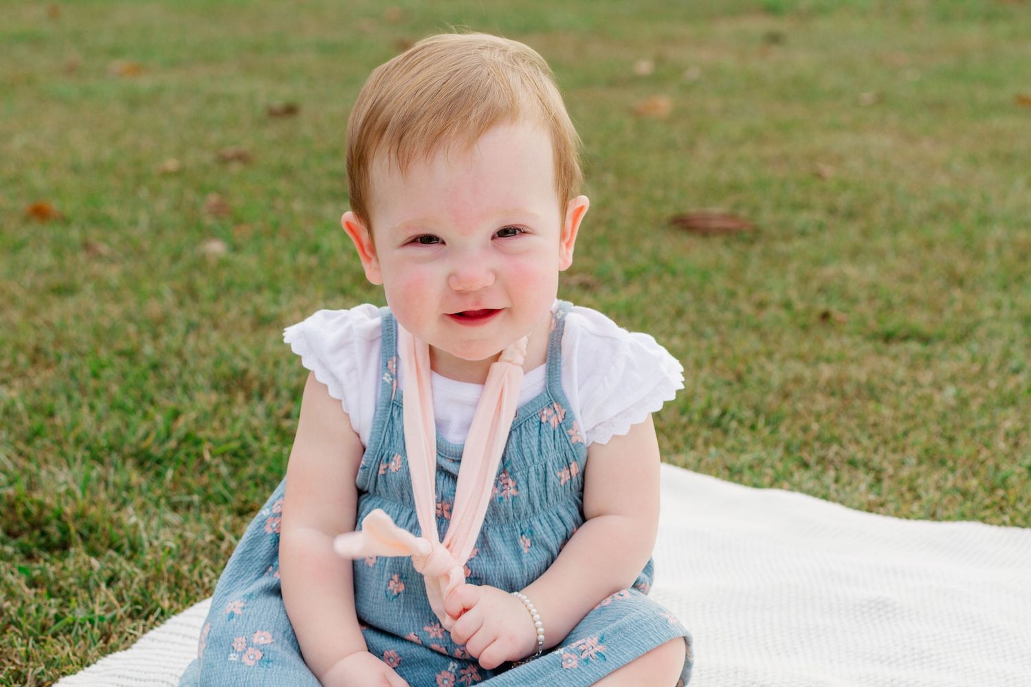 A baby in a blue dress sits on a white blanket in the grass during an outdoor photo session.