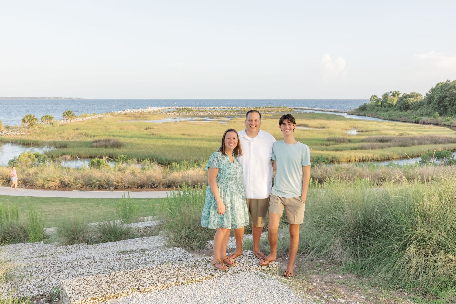 Family of three posing on a gravel path surrounded by marsh grasses with a river and blue sky behind.