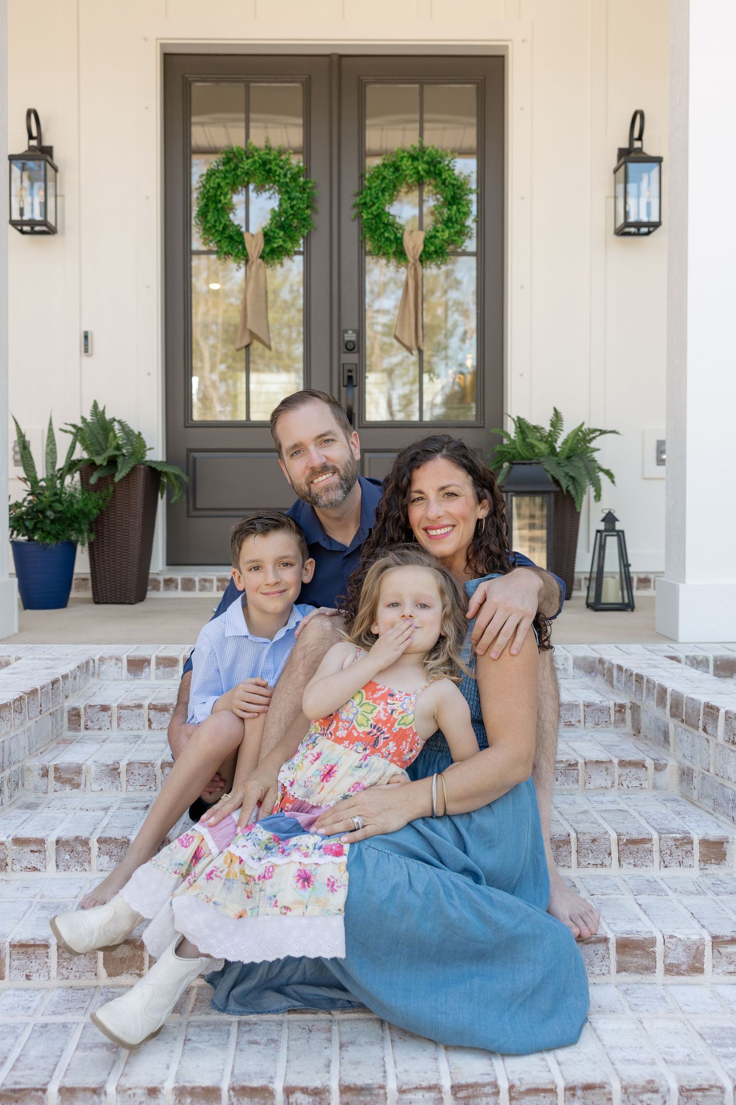 Happy family of four posing on brick front porch steps of a white home with green wreaths on double doors.