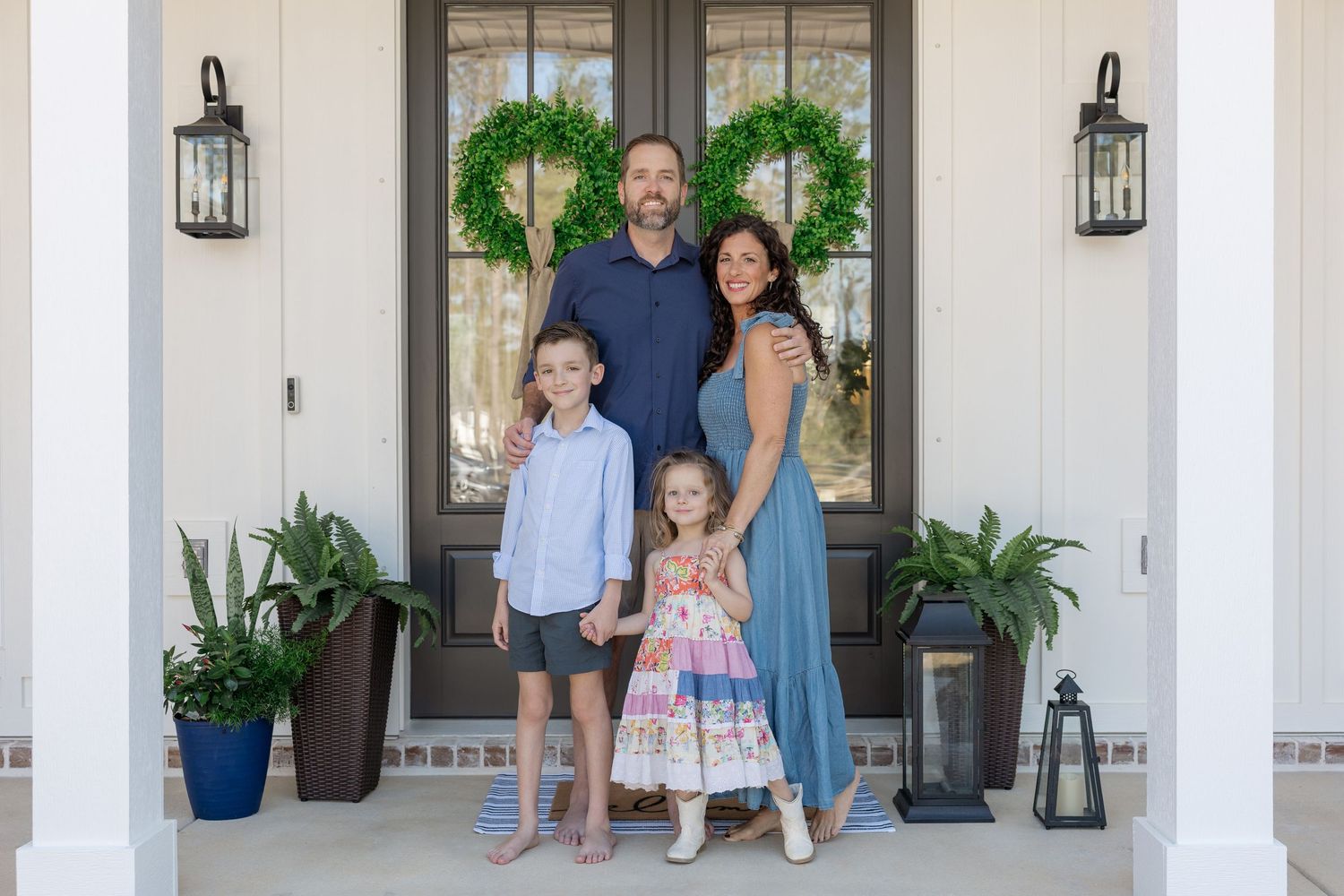 Family of four posing on a welcoming front porch with white columns, green wreaths, and lanterns.