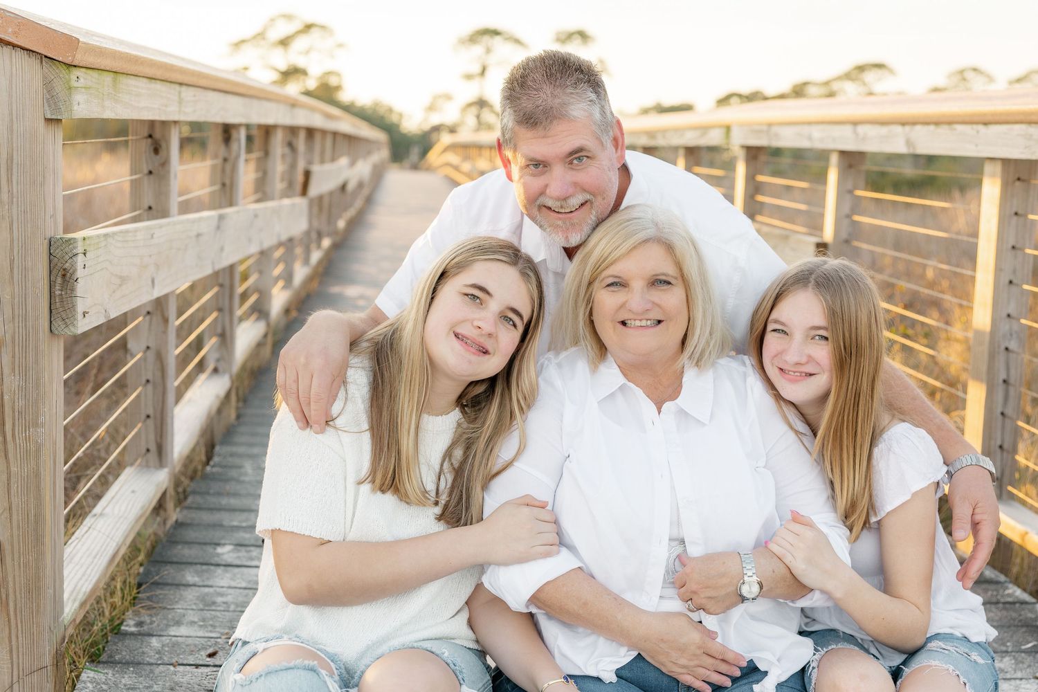 A family wearing white sits together on a wooden boardwalk during sunset, creating a warm and intimate portrait.