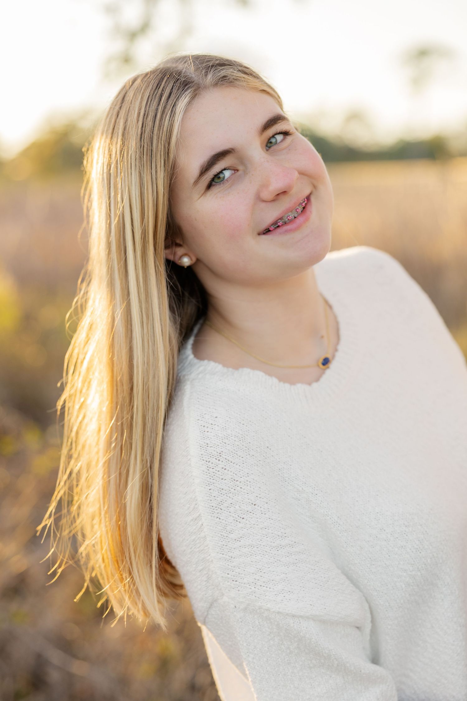 A person in a white sweater poses outdoors during golden hour with natural backlighting.