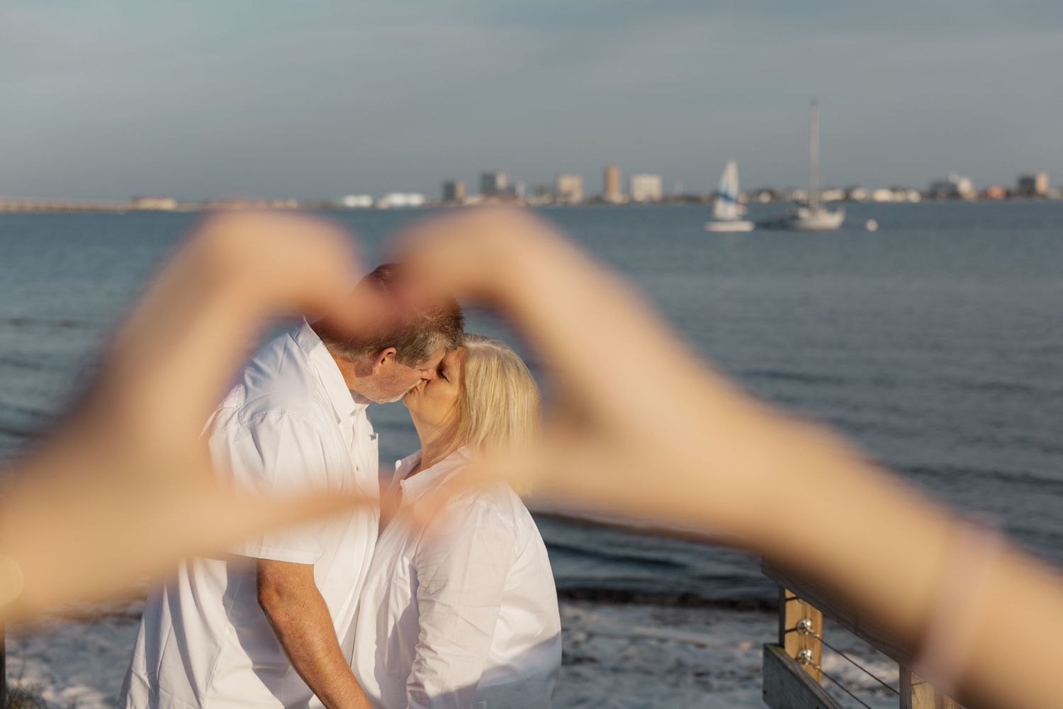 Two people share a romantic moment by the water as hands form a heart shape in the foreground with boats visible in the distance.