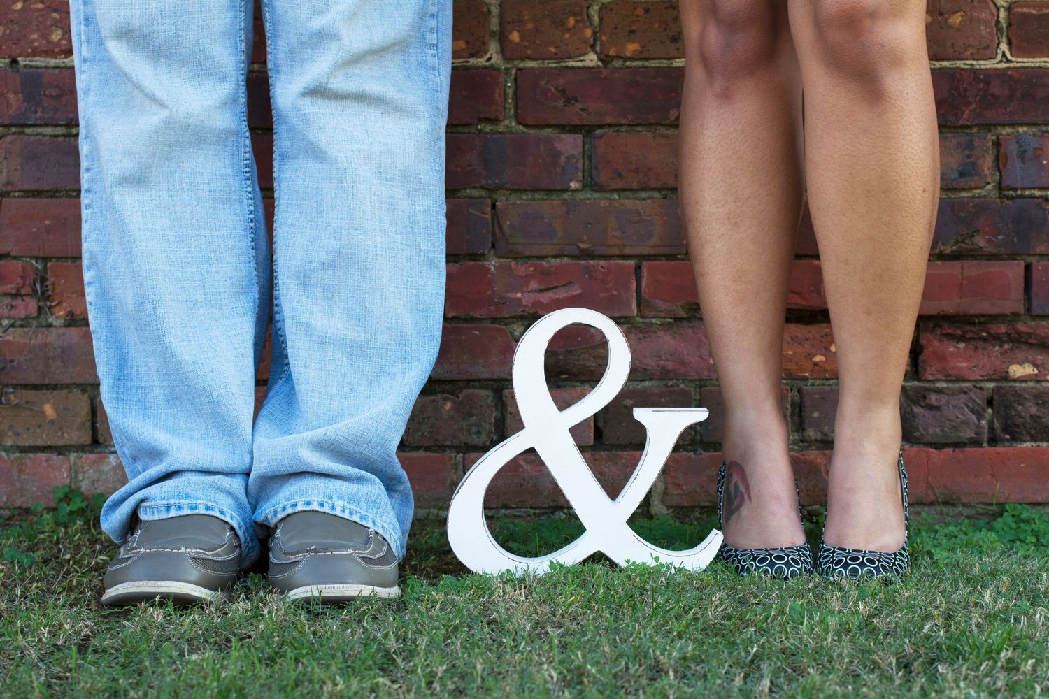 A row of people's legs and feet shown against a brick wall background.