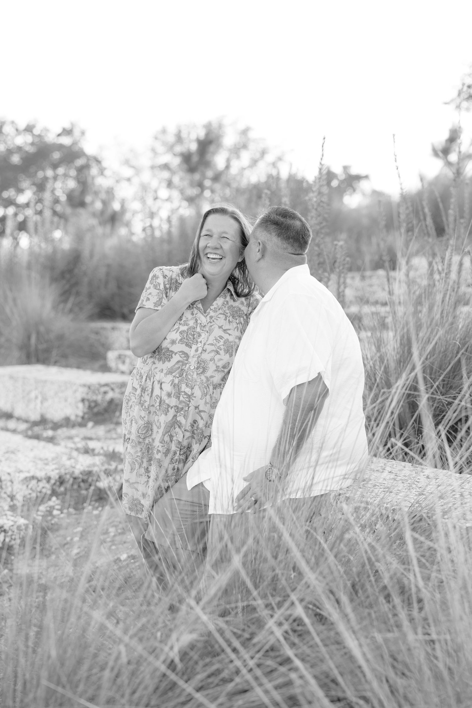 Black and white photo of a laughing couple sharing a joyful moment outdoors among tall grasses and rocks.