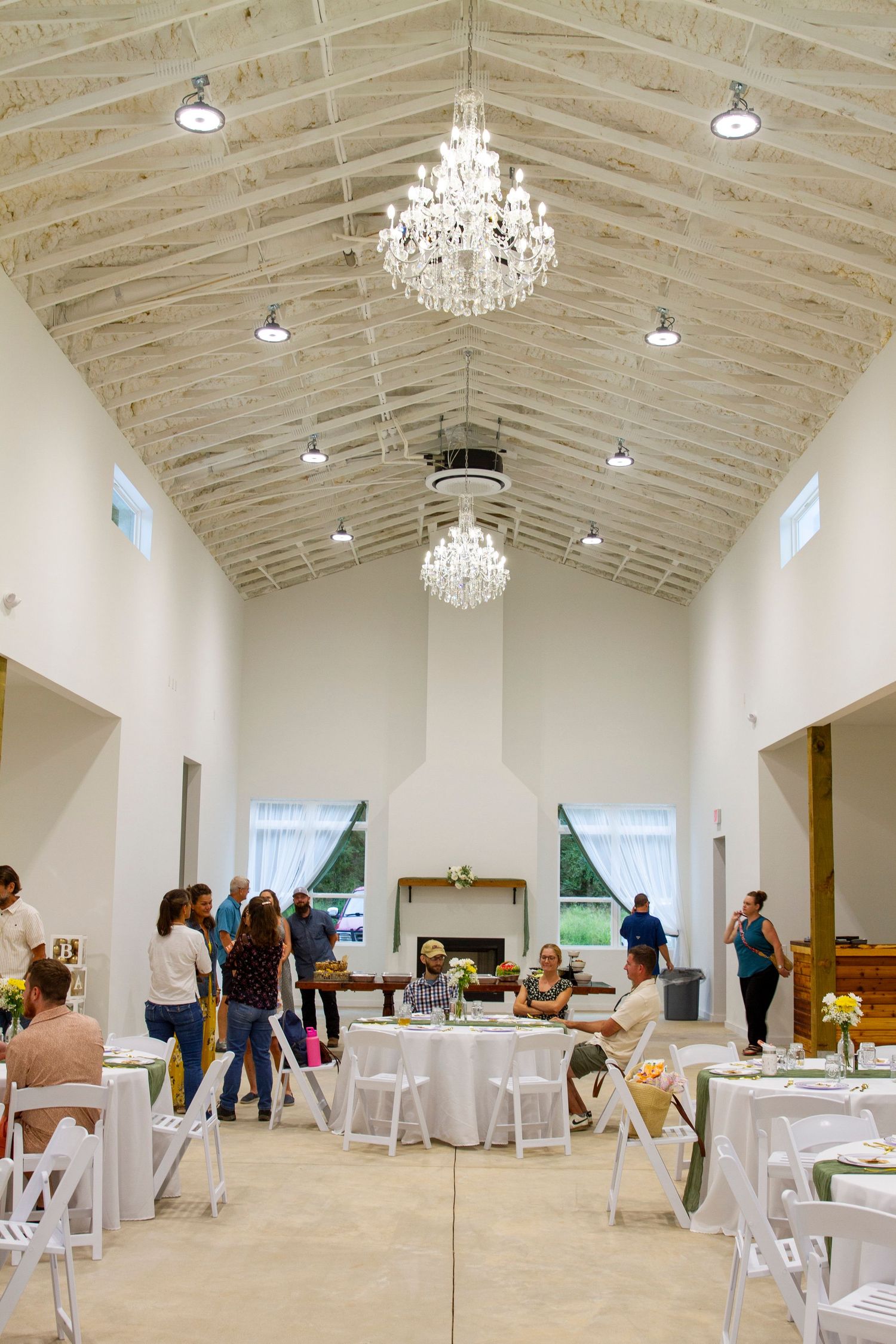 A modern event venue with vaulted white ceilings, crystal chandeliers, and round tables set up for a gathering.