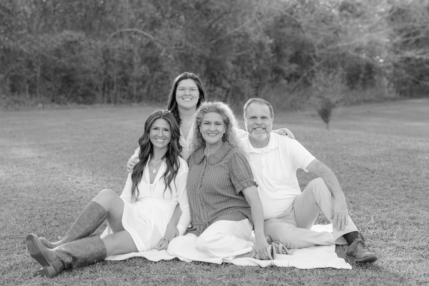 Black and white family portrait of four people smiling outdoors on grass with trees in background.