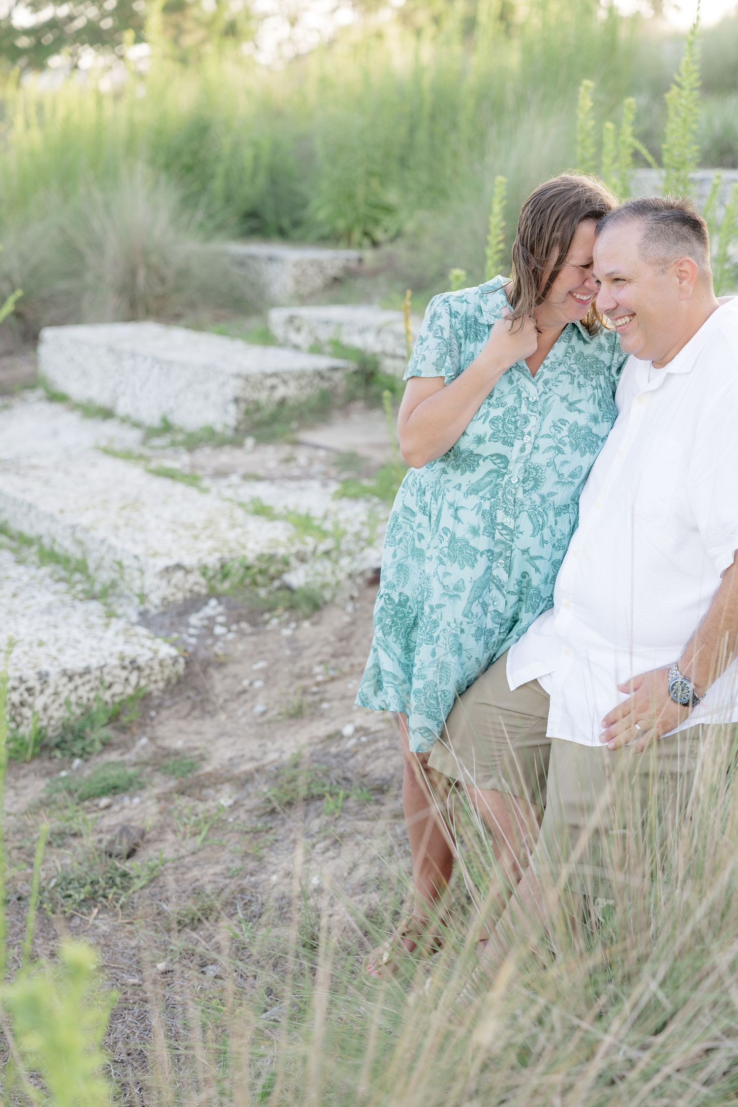 Couple laughing together outdoors near stone steps surrounded by lush green plants during a romantic engagement photo session.