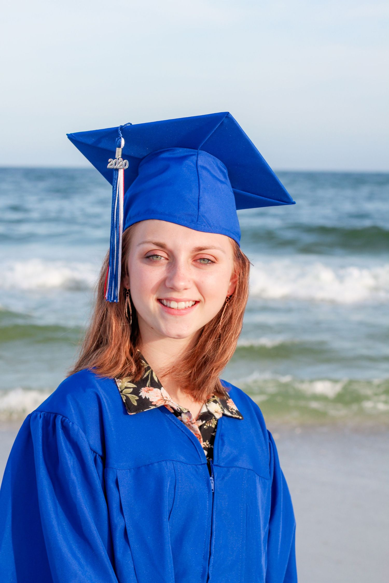 Graduate in bright blue cap and gown standing on beach with ocean waves in background.