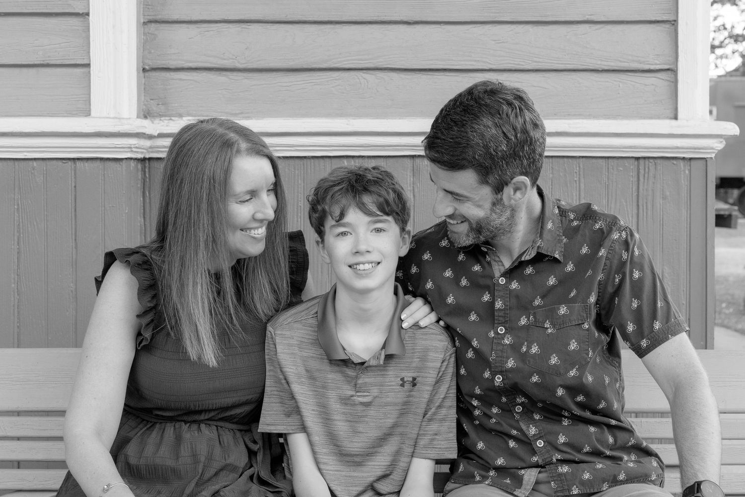 Black and white portrait of a smiling family of three sitting together on a porch bench.