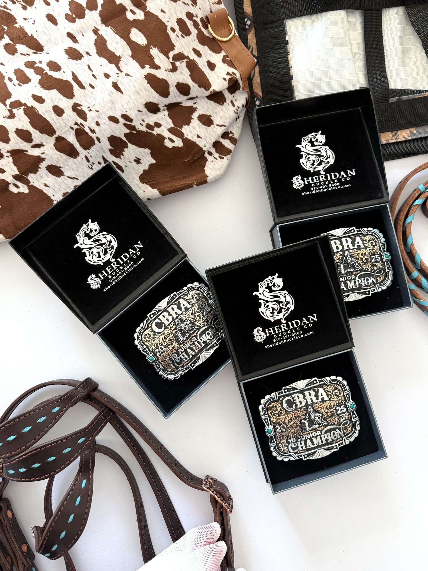 Branded black jewelry boxes with silver belt buckle designs displayed alongside brown leather straps on cowhide print background.