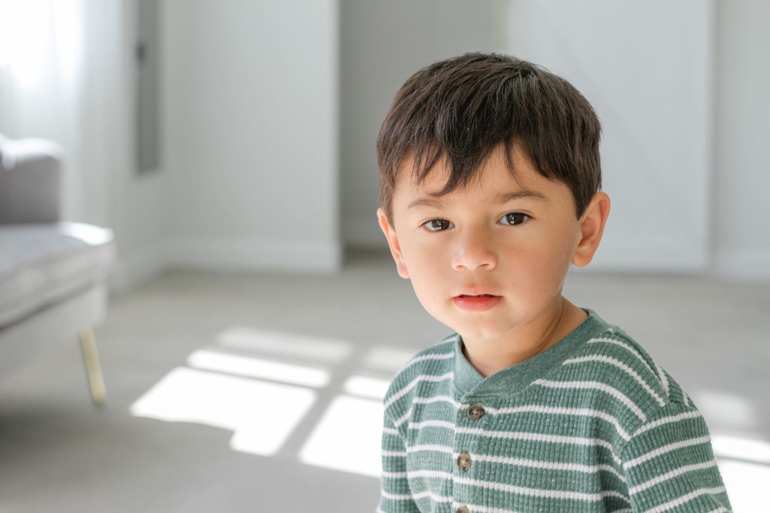 A small child wearing a green and white striped shirt stands in a bright, sunlit room.
