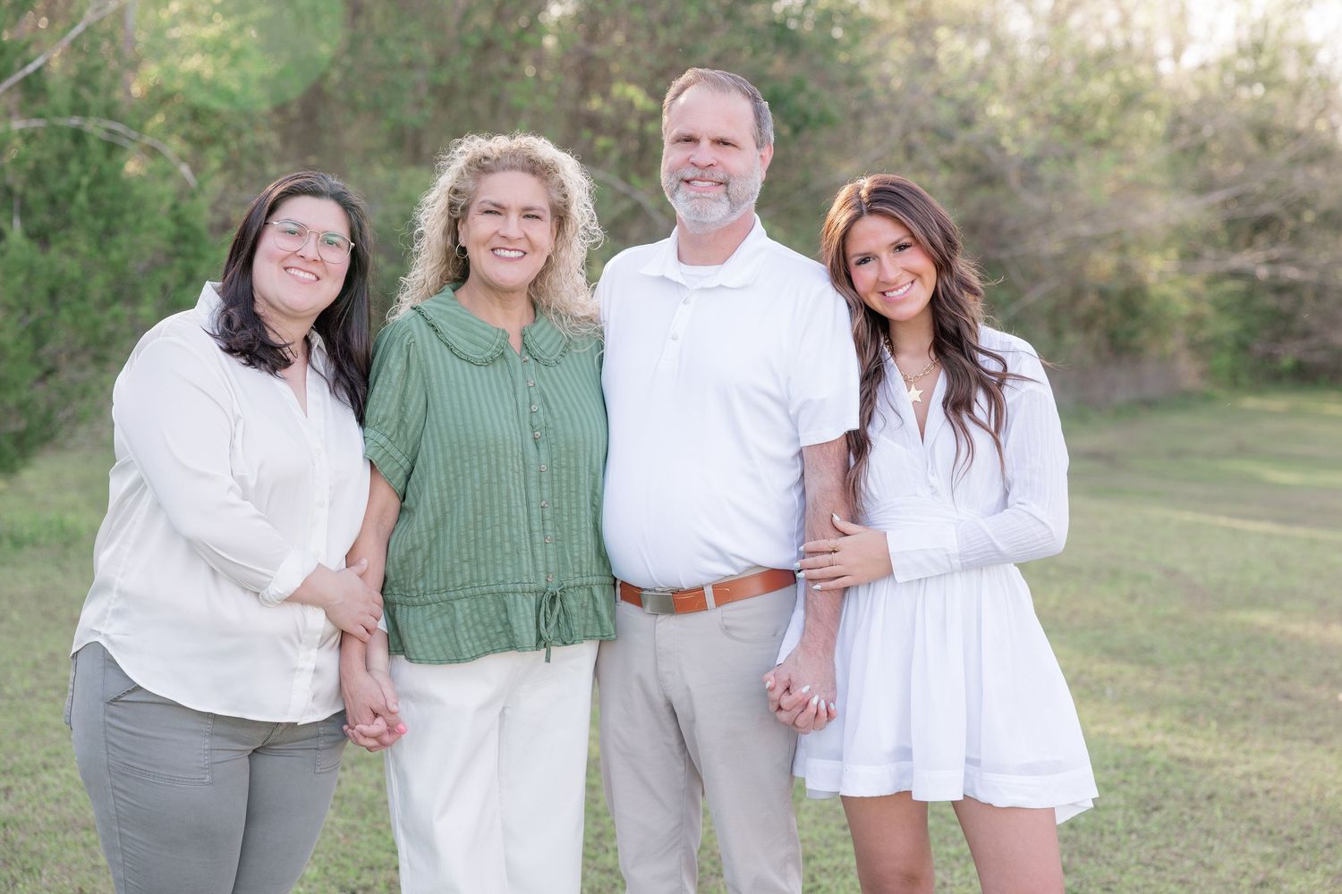 Family of four smiling outdoors in a green park, dressed in white and green casual attire for a portrait session.