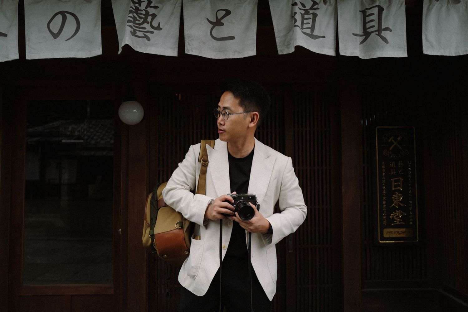 A person poses in front of Japanese storefront signage wearing a white blazer and black shirt.