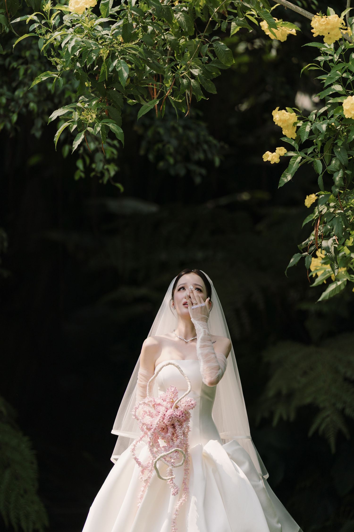 Bride in white gown and veil holds pink bouquet under yellow flowering trees in a lush garden setting.