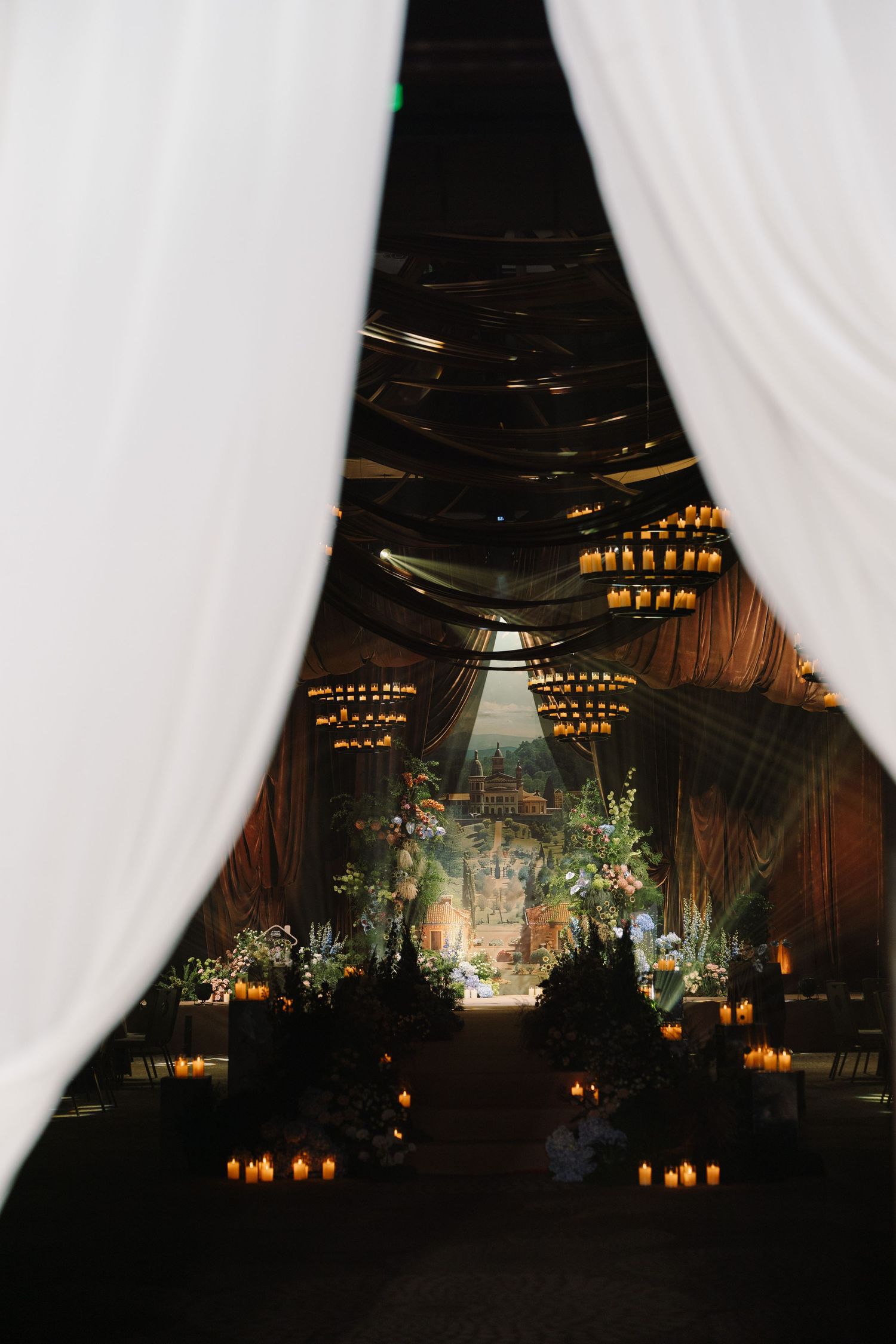Elegant wedding venue with white draping, candlelit aisle, floral arrangements, and chandelier lighting in a dark romantic setting.