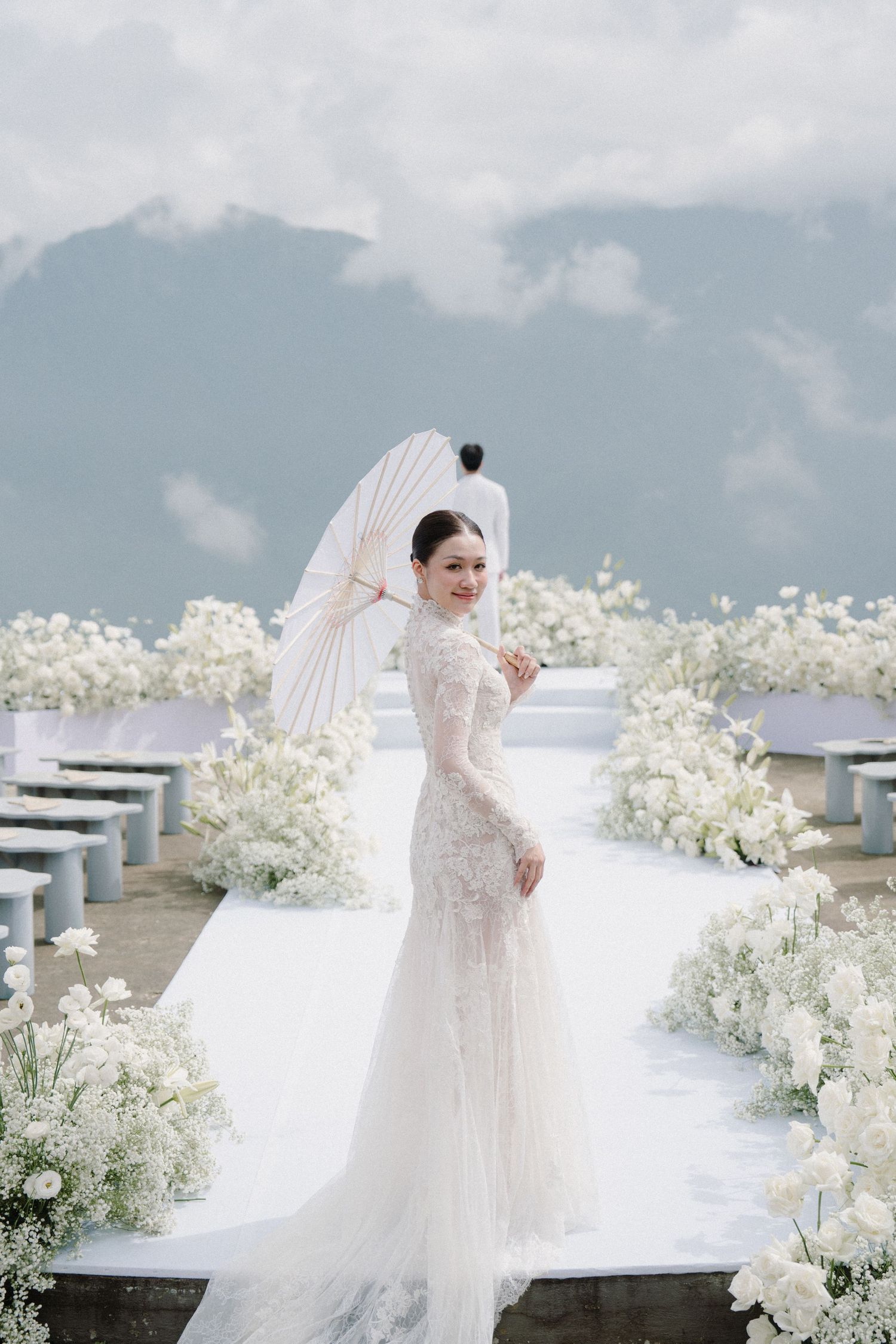 A bride in a flowing white dress poses with white floral arrangements in an elegant cloudy outdoor setting.