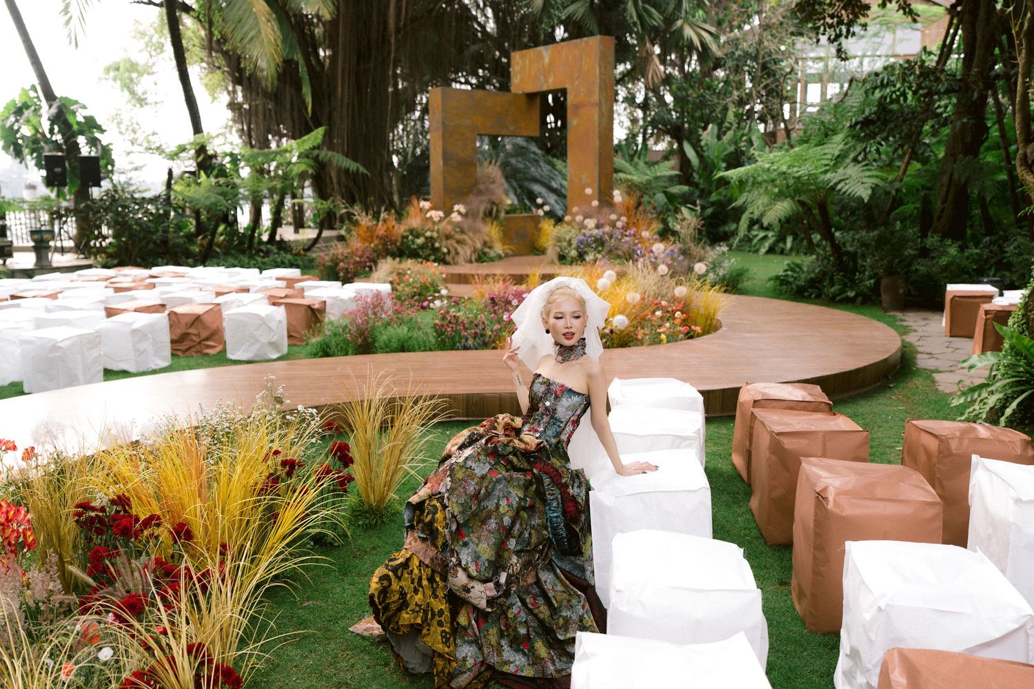 A young girl in a floral gown poses at an outdoor garden event with white chairs and lush tropical greenery.