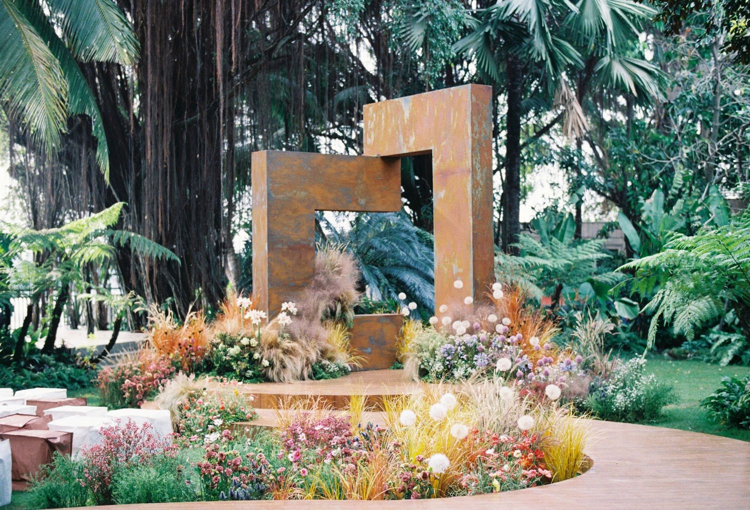 Modern geometric metal sculpture surrounded by colorful wildflowers in a lush tropical garden setting.