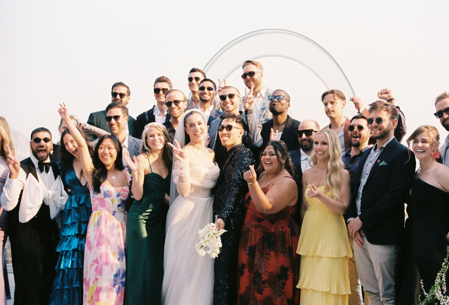 A panoramic group photo of wedding guests wearing sunglasses and formal attire standing in a line outdoors.