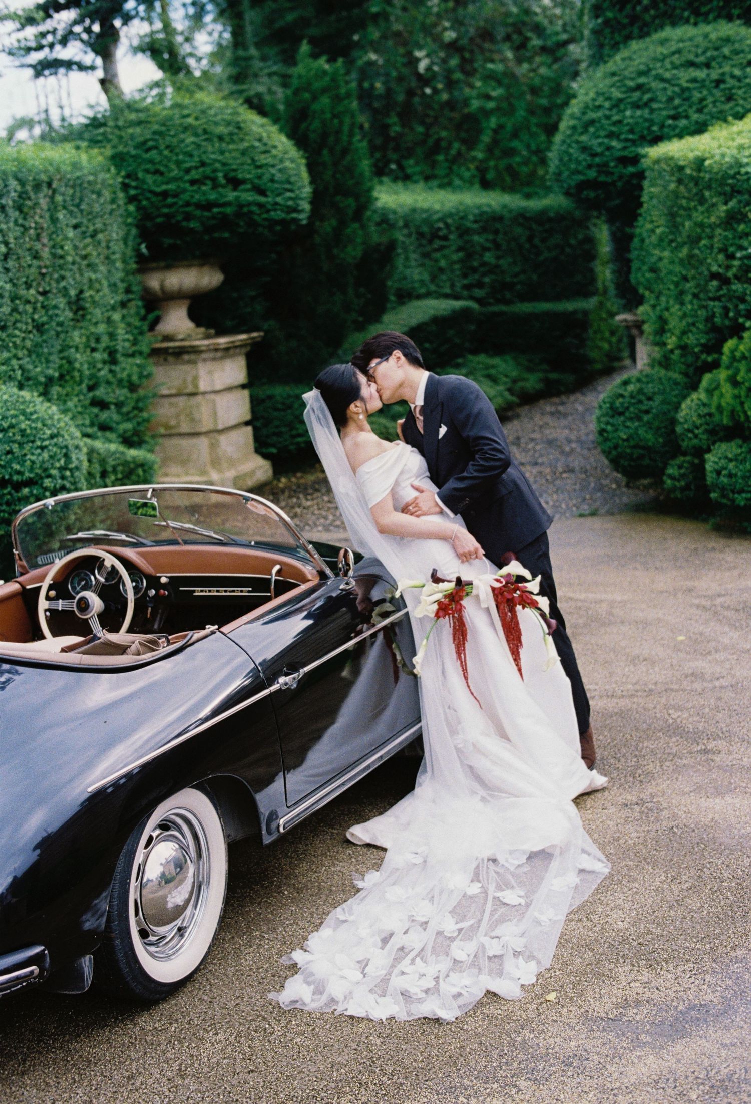 A romantic wedding scene with a vintage black convertible car parked in a formal garden with manicured hedges.