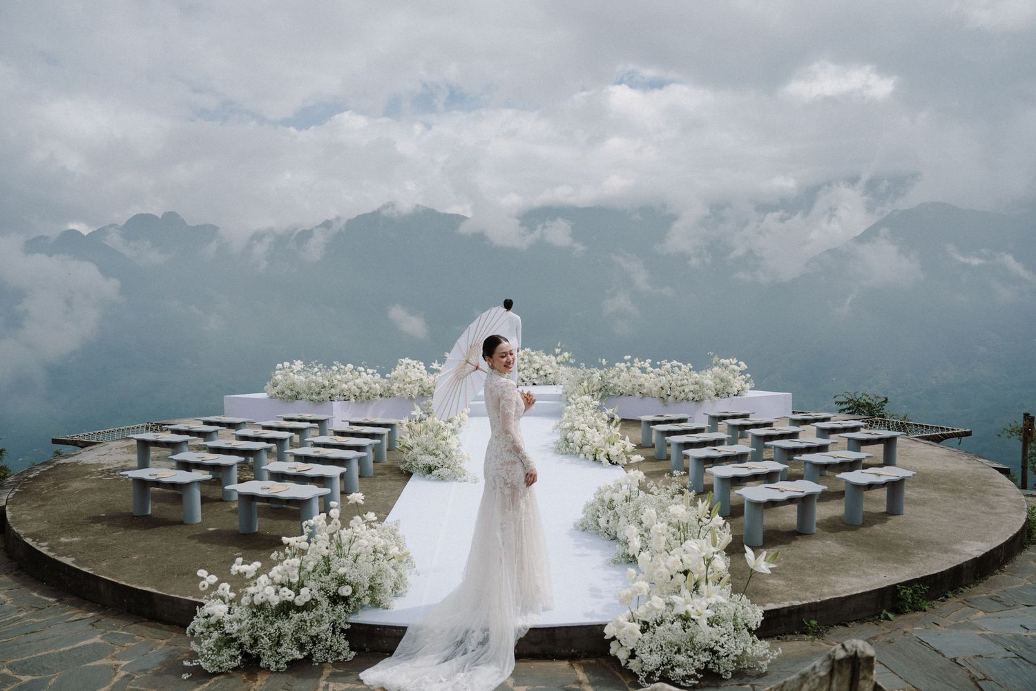 A couple in wedding attire stands on a scenic mountain overlook surrounded by benches and white floral arrangements.