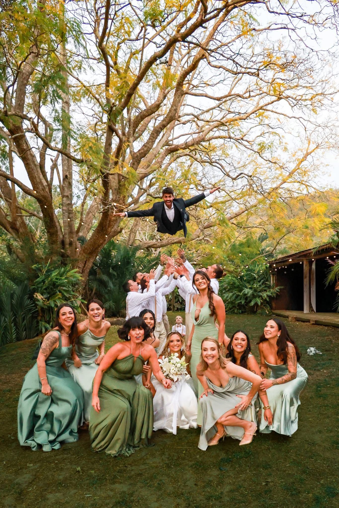 Wedding party in sage green dresses posing playfully on grass under autumn trees.