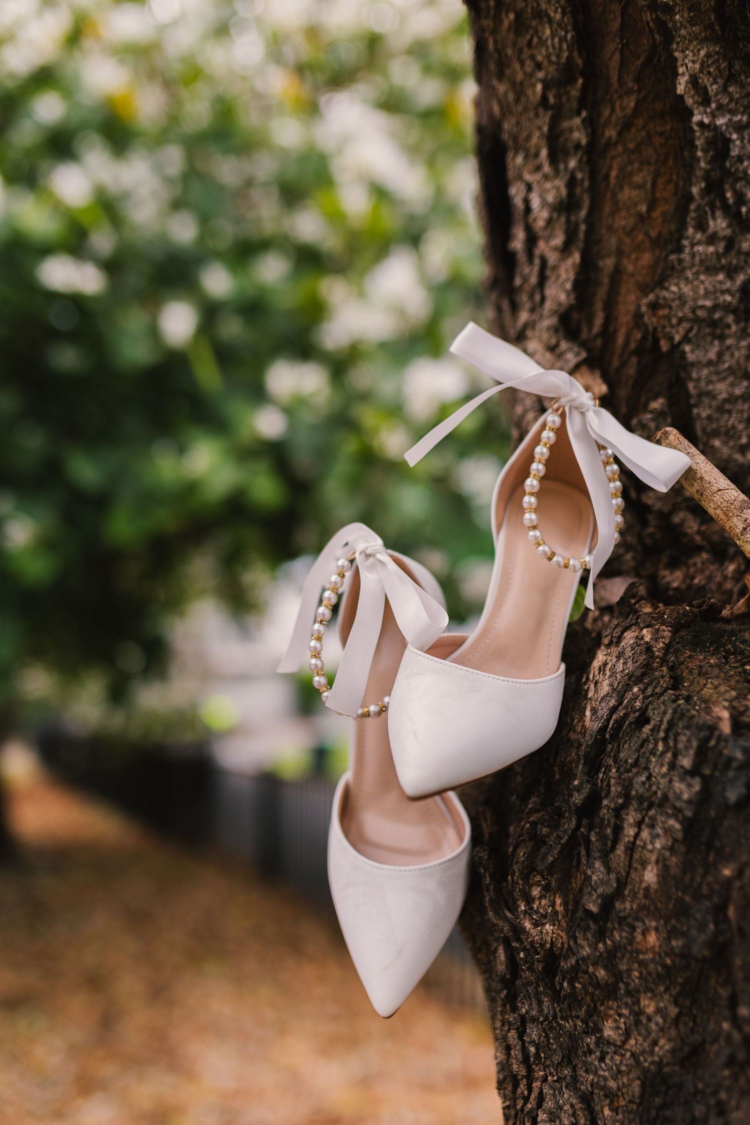 Elegant white bridal heels with ankle straps and pearl details displayed against a tree trunk in a garden setting.