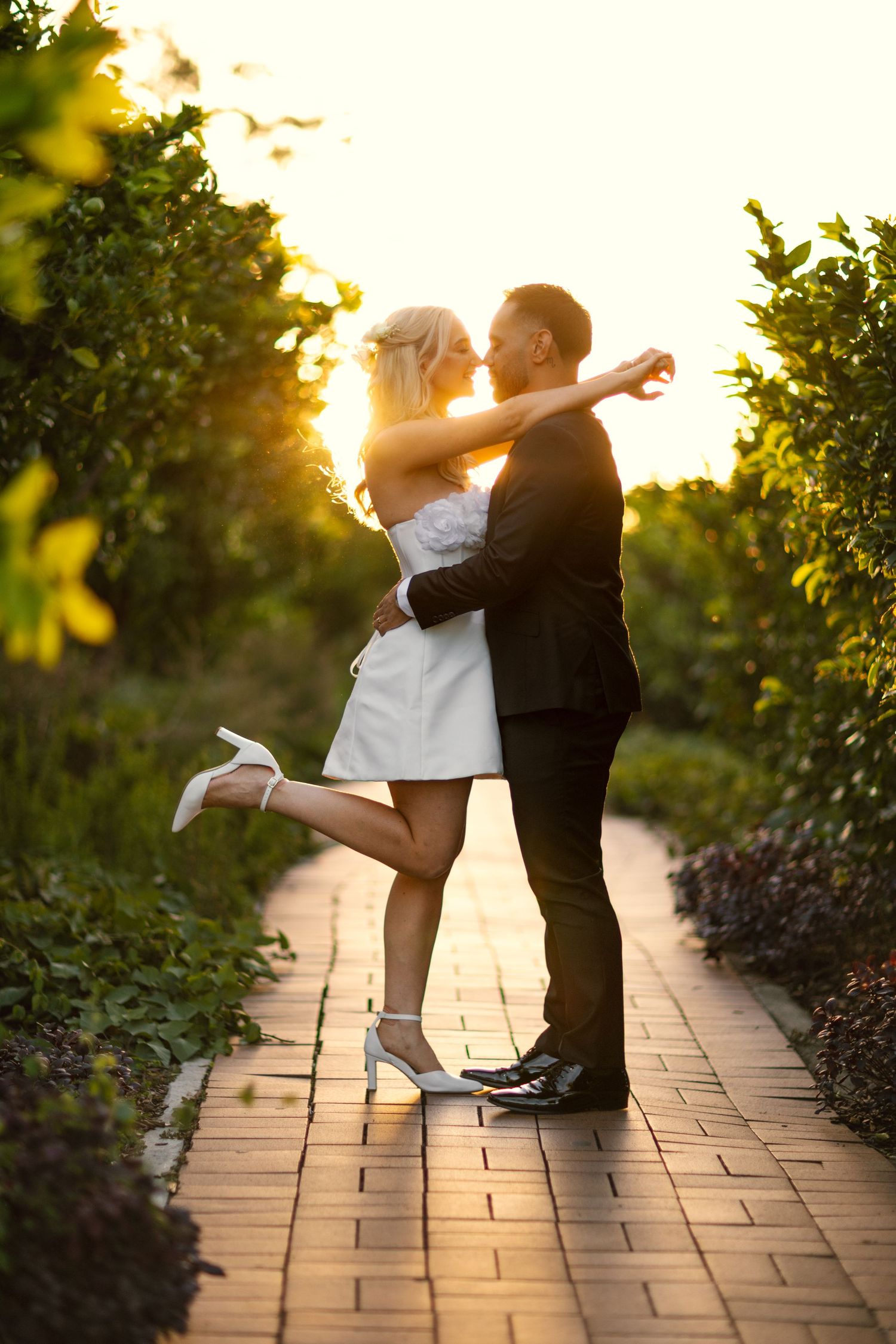 A romantic sunset silhouette of a couple embracing on a brick pathway surrounded by lush garden foliage.