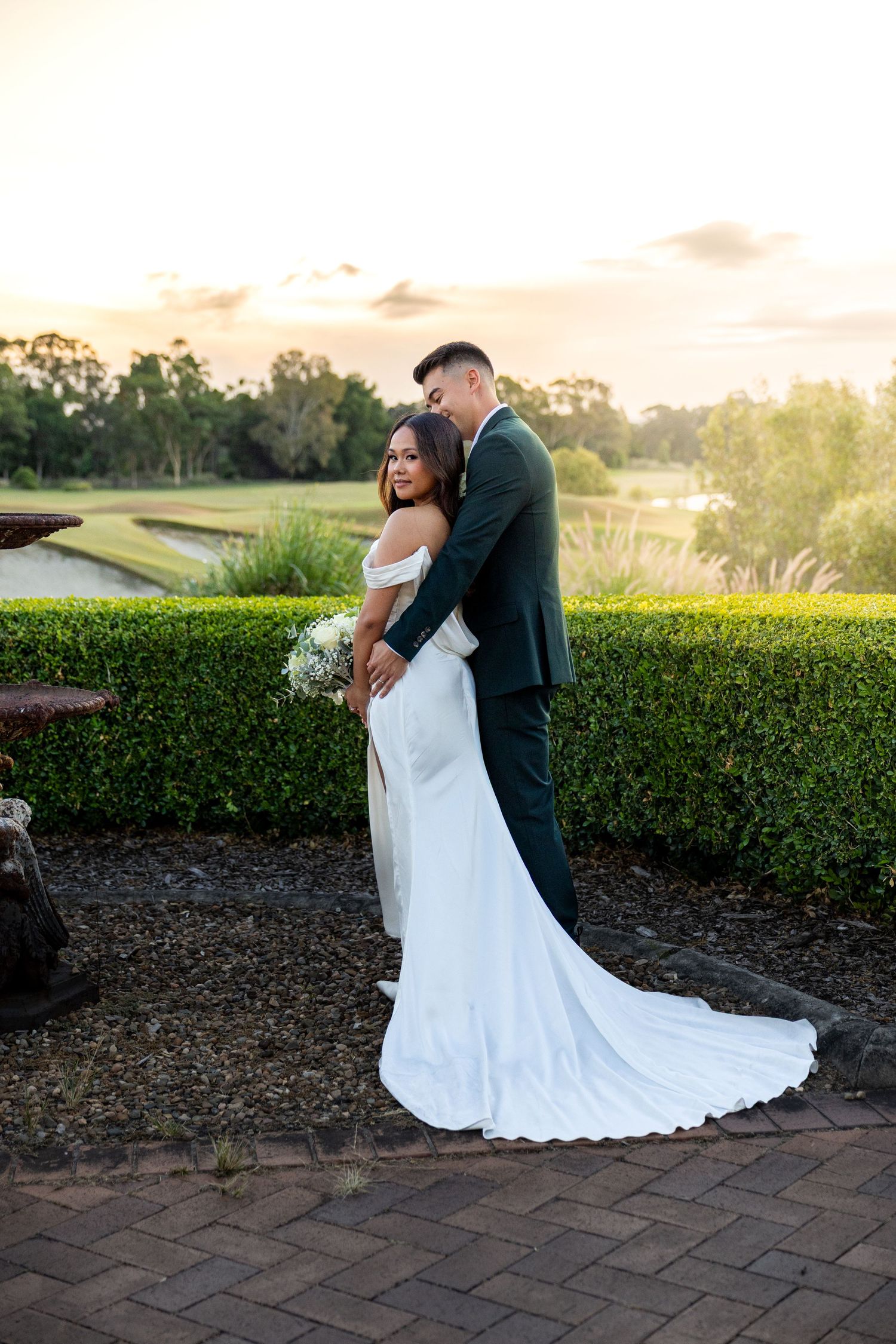 A couple embraces during golden hour at a garden venue with manicured hedges and brick pathways.