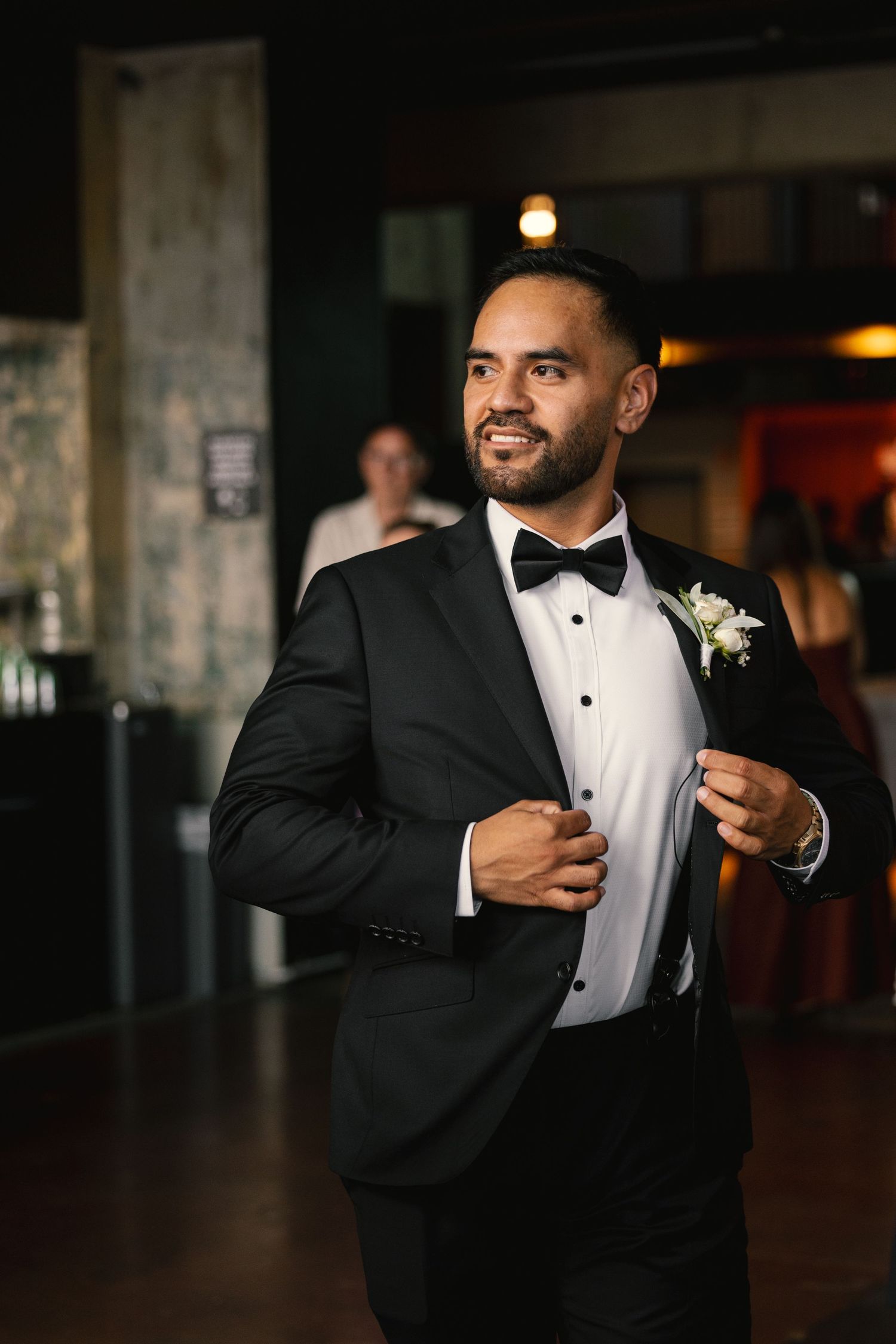 Groom in classic black tuxedo and bow tie smiles while adjusting his jacket.