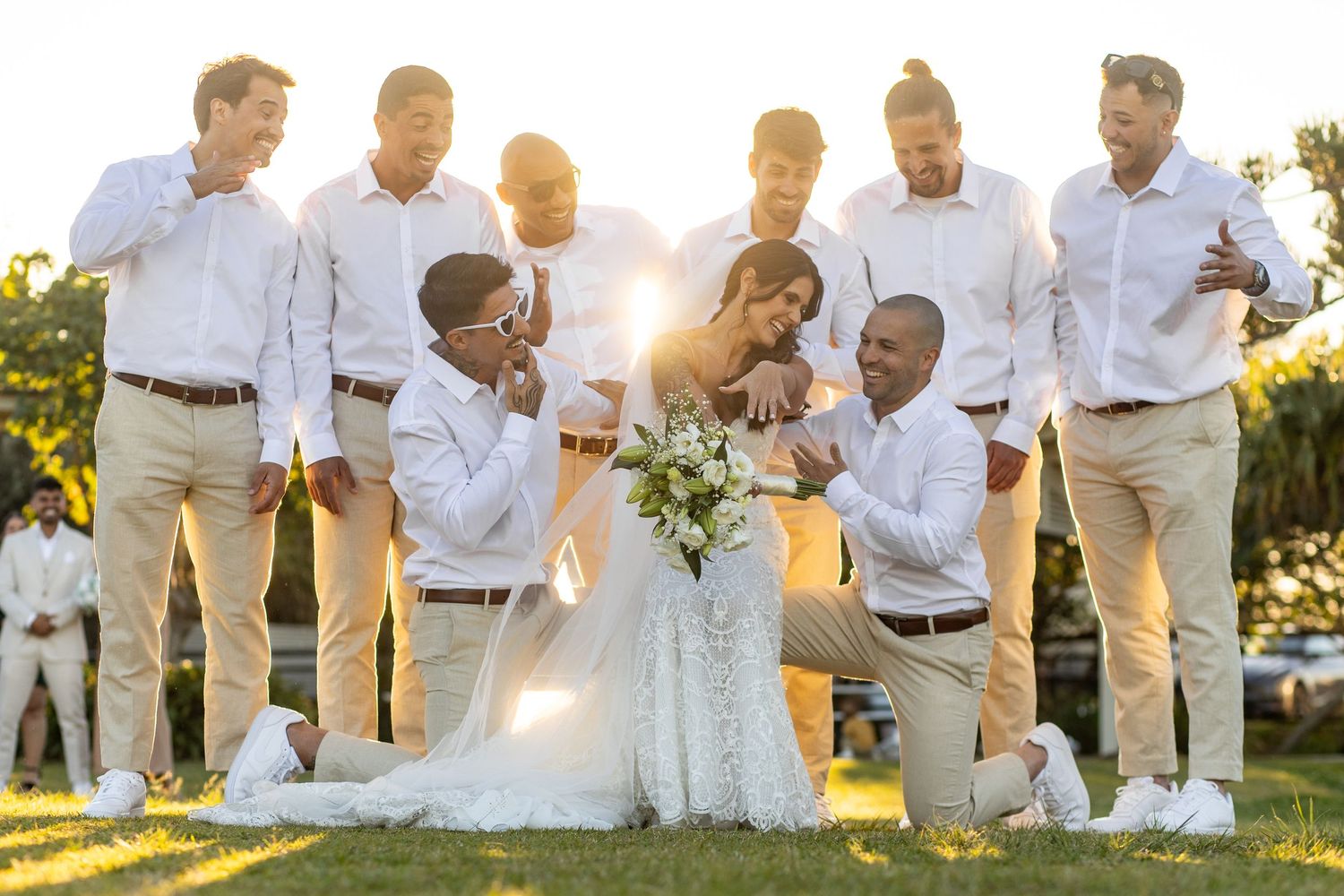 Wedding party in white shirts and khaki pants gather on sunny lawn during golden hour celebration.